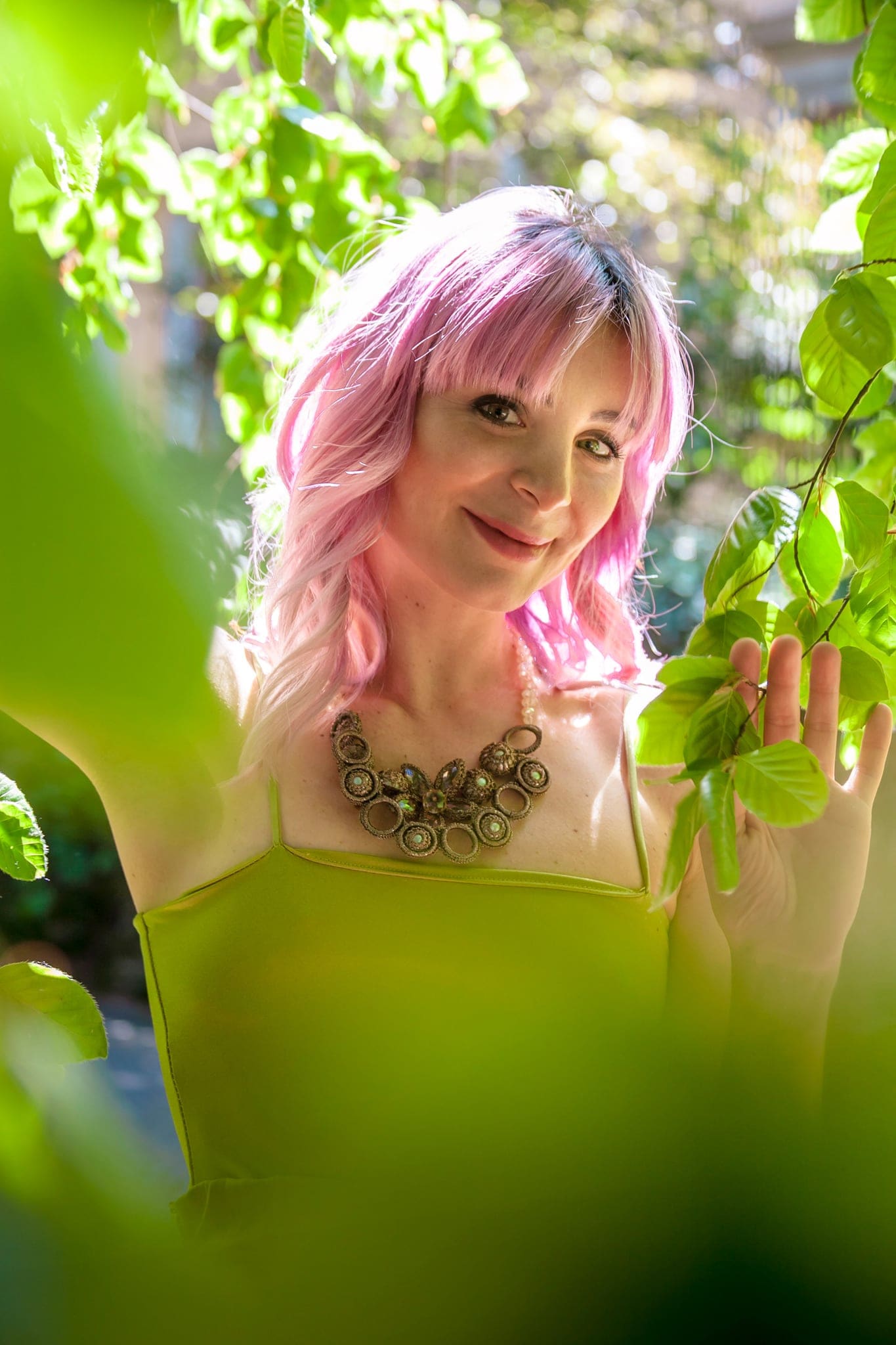 Donna dai capelli rosa con collana, sorridente tra le foglie verdi di un albero, in un ritratto all'aperto dai toni luminosi