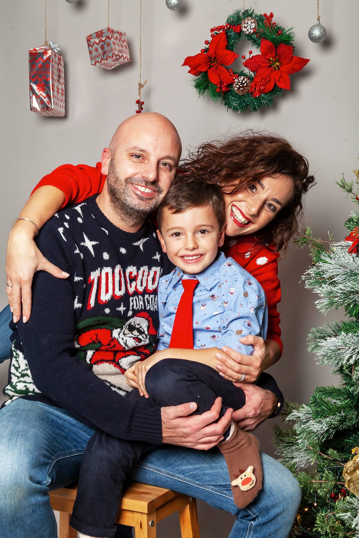 Papà, mamma e figlio con camicia azzurra e cravatta rossa, seduti su uno sgabello con ghirlanda e regali appesi alle spalle
