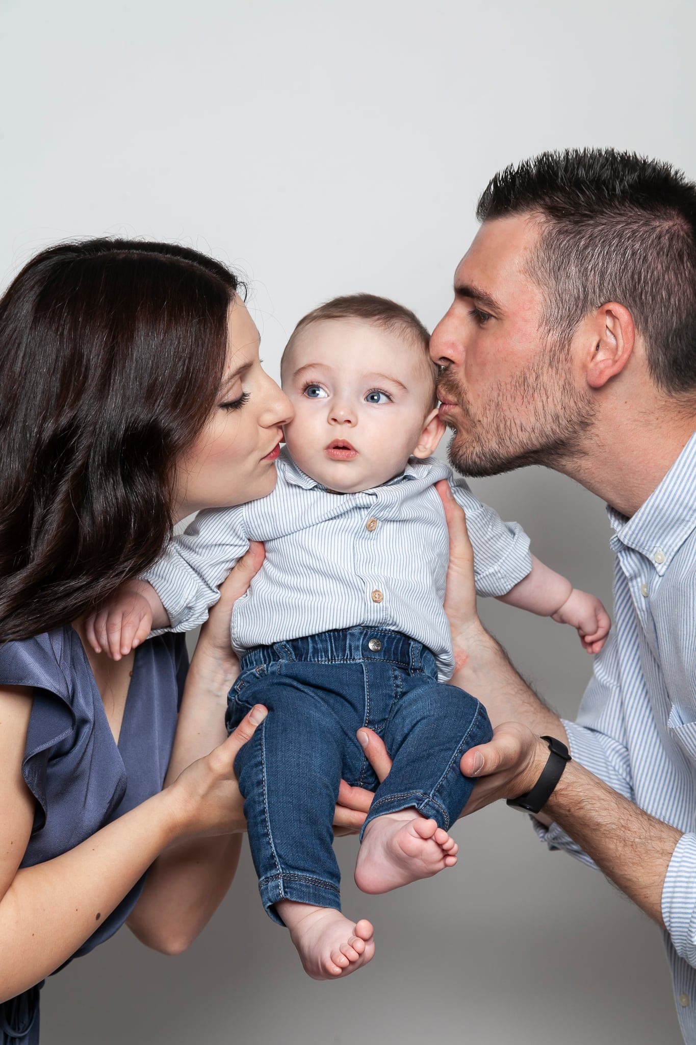 Mamma e papà baciano le guance del loro bimbo in camicia azzurra e jeans, tenuto in alto su sfondo grigio