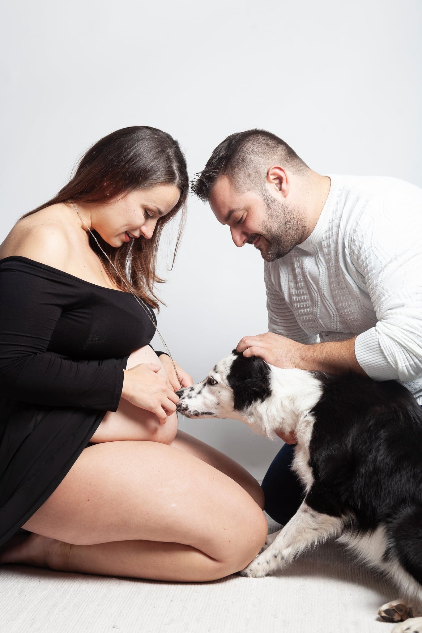 Coppia inginocchiata fronte contro fronte con il loro cane bianco e nero che annusa il pancione, in uno shooting di gravidanza in studio.
