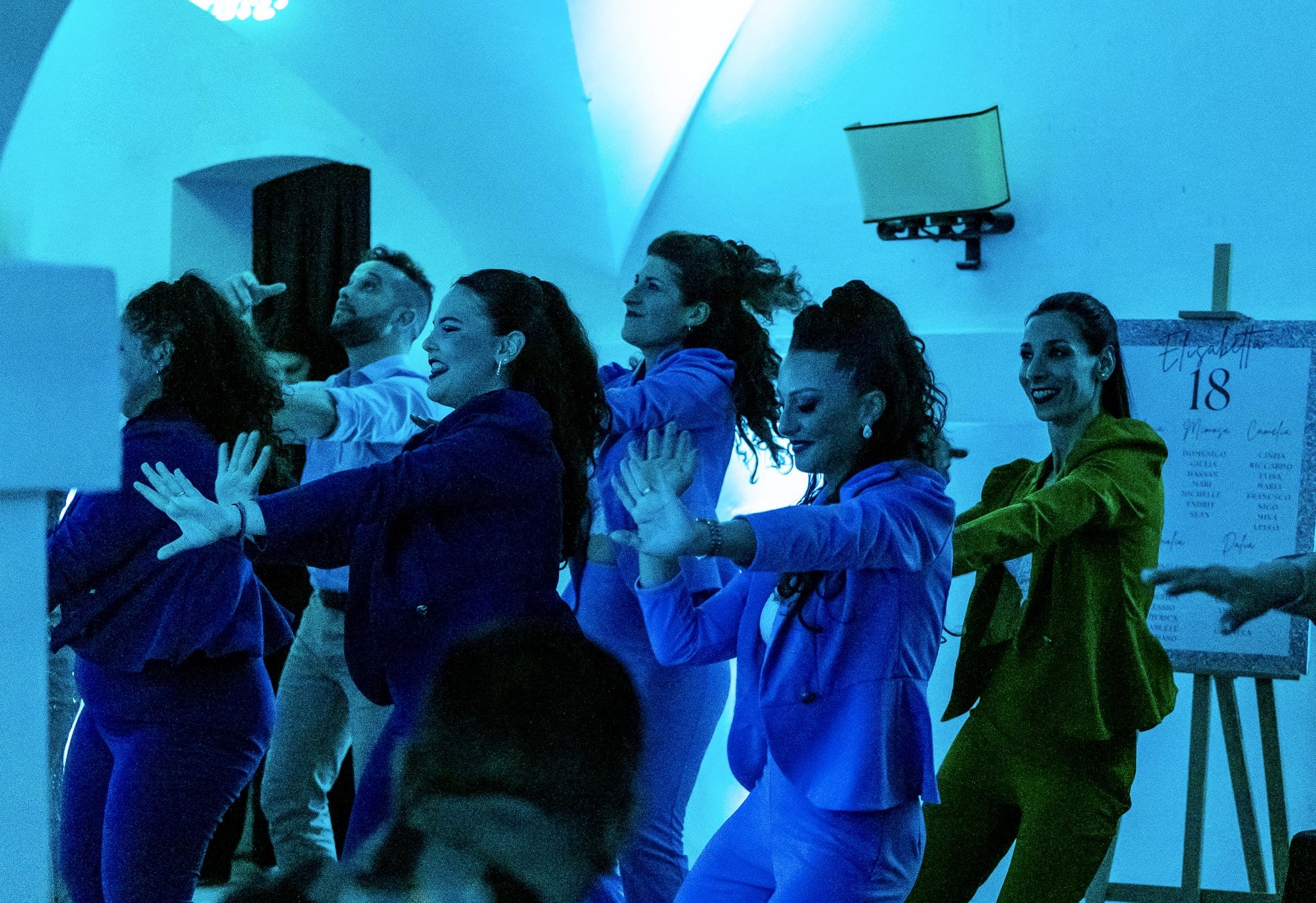 Invitate in giacche blu, viola e verdi ballano con le braccia aperte sotto luci azzurre, accanto al cartellone dei tavoli