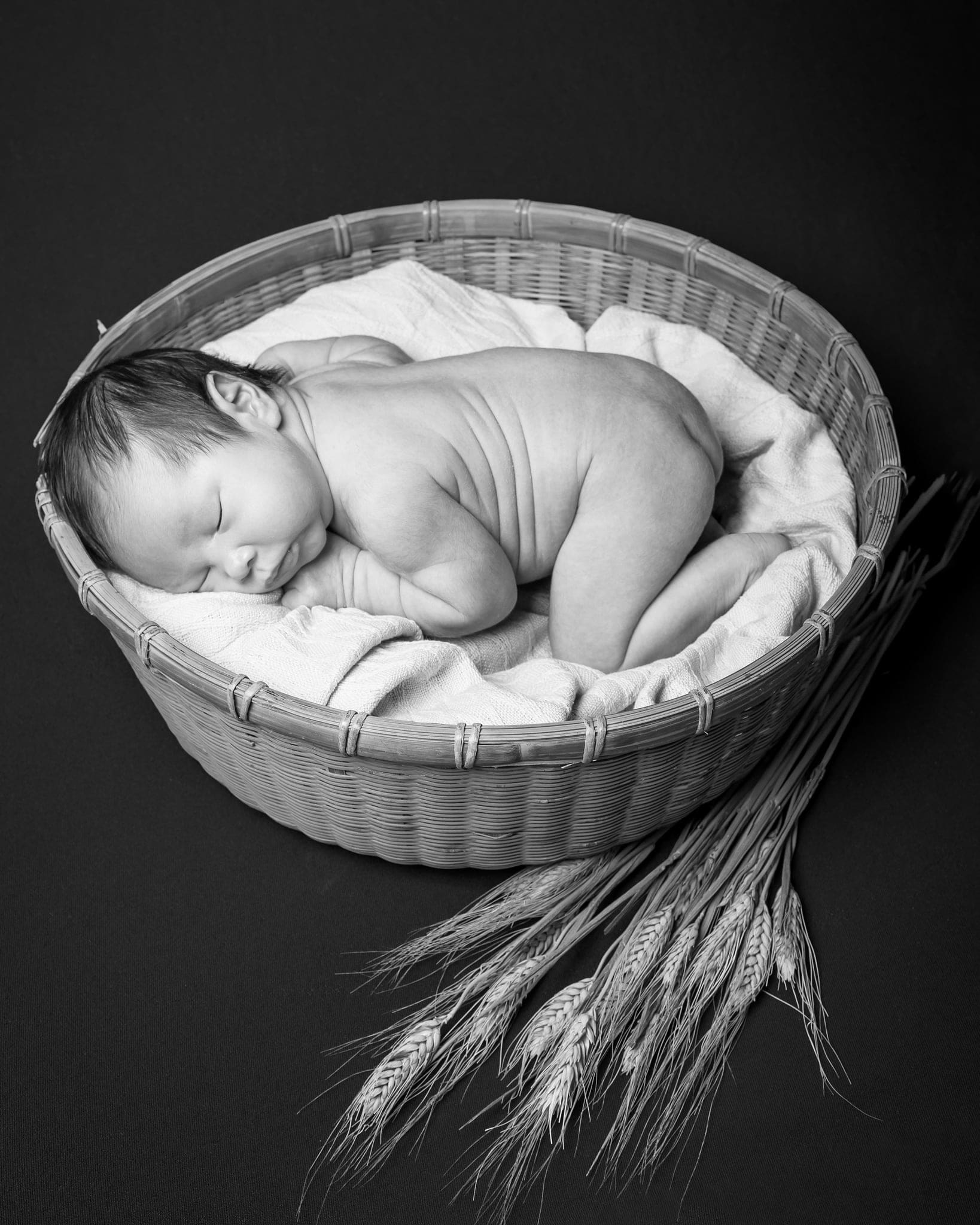 Neonato che dorme raggomitolato in un cesto con una spiga di grano accanto, in bianco e nero su fondo scuro.