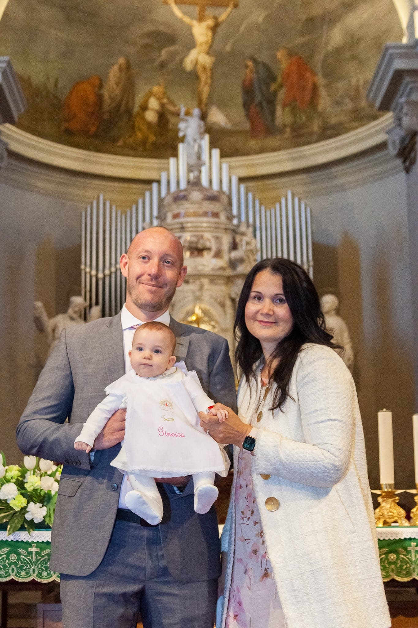 Ritratto davanti all'altare di chiesa: papà e mamma sorridono tenendo la bimba in abito bianco, con le canne dell'organo dietro