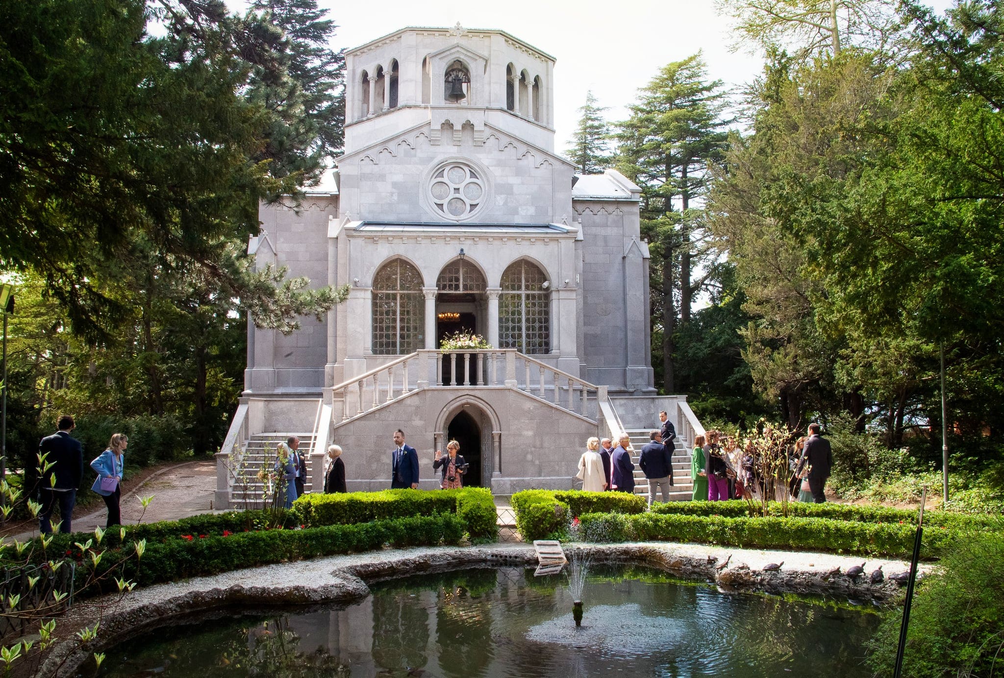 Cappella in pietra chiara nel verde con piccolo laghetto e invitati radunati davanti, matrimonio al Parco Revoltella di Trieste