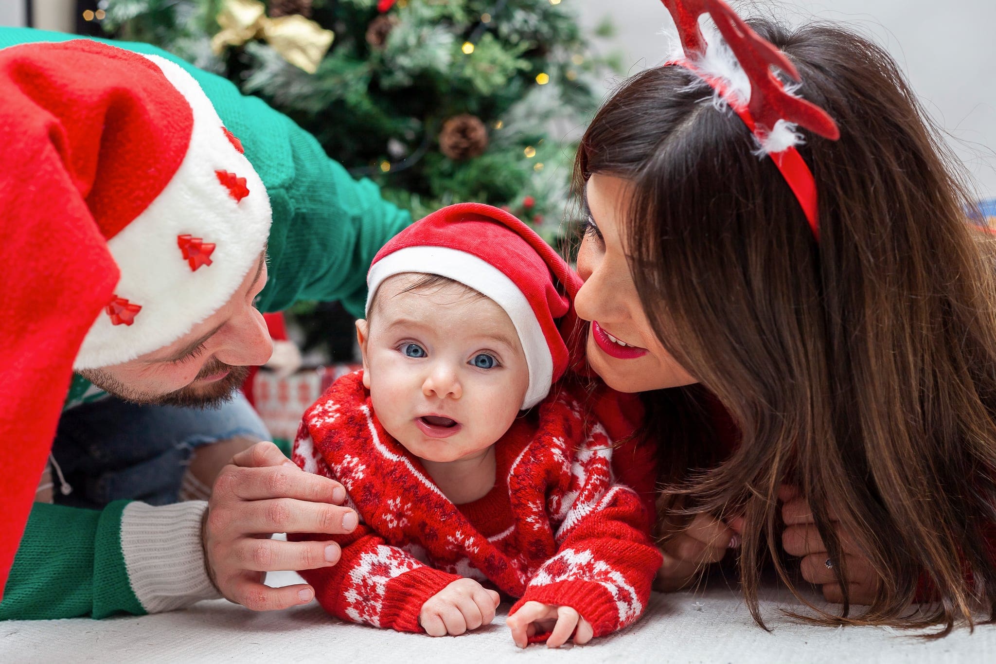 Mamma e papà sdraiati a terra con cappello di Babbo Natale e cerchietto a renna guardano il neonato con tutina rossa