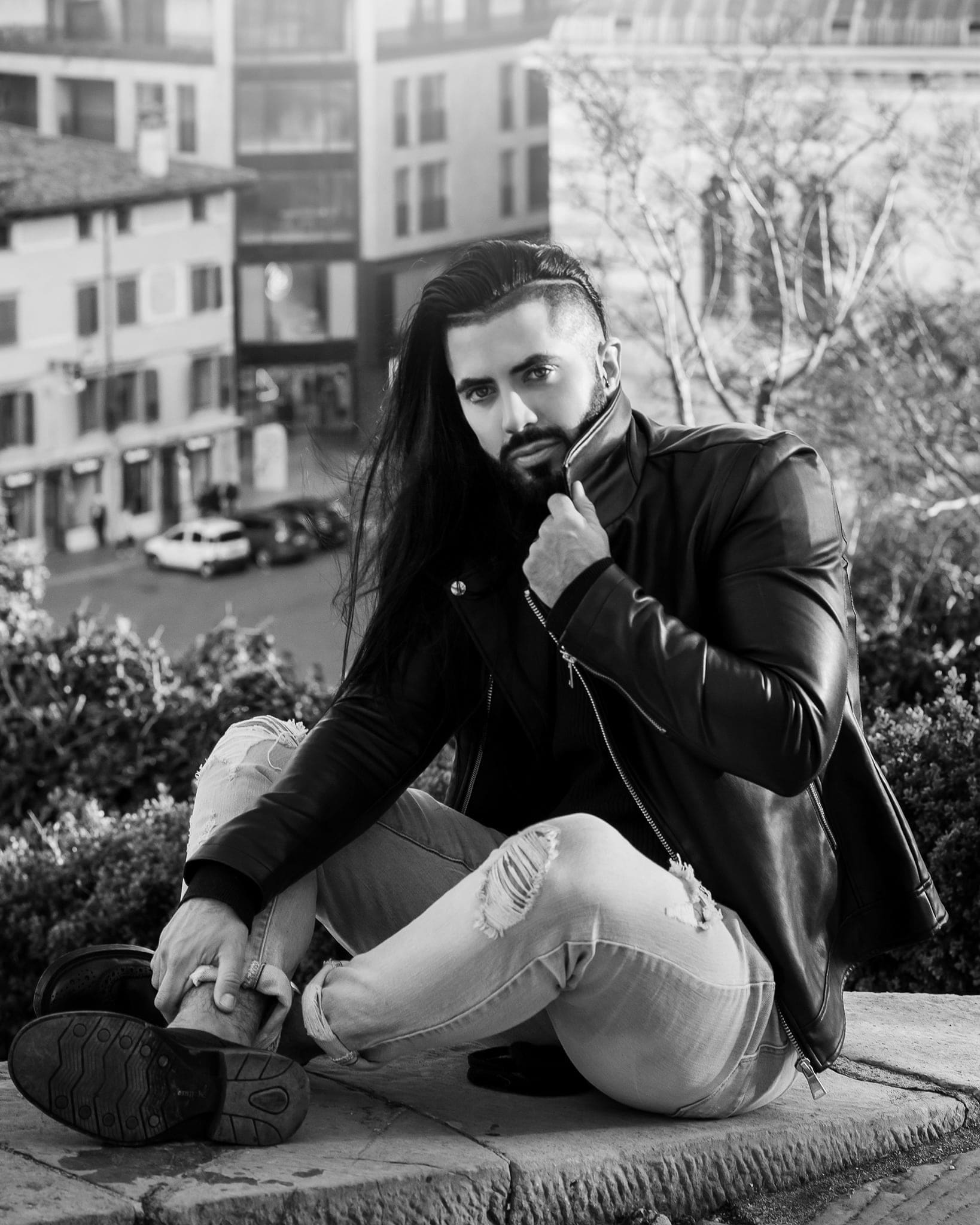 In bianco e nero, uomo con lunghi capelli neri e barba, in giacca di pelle e jeans strappati, seduto su un muretto con edifici sullo sfondo