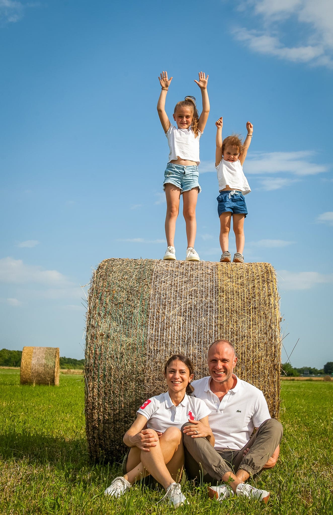 Due bambine con le braccia alzate in piedi sopra una balla di fieno, mentre mamma e papà siedono sotto, cielo azzurro