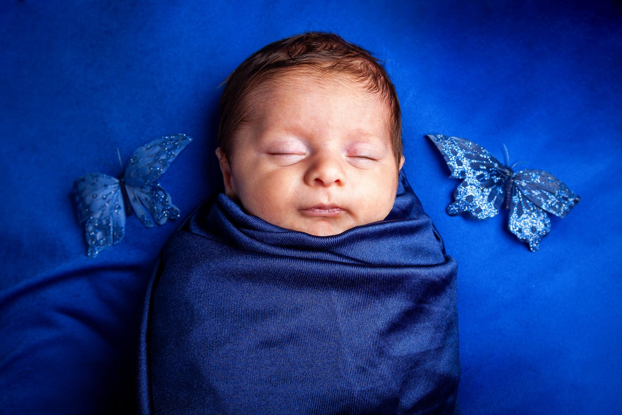 Neonato che dorme avvolto in un telo blu scuro, con due farfalle decorative blu ai lati, su fondo blu elettrico.