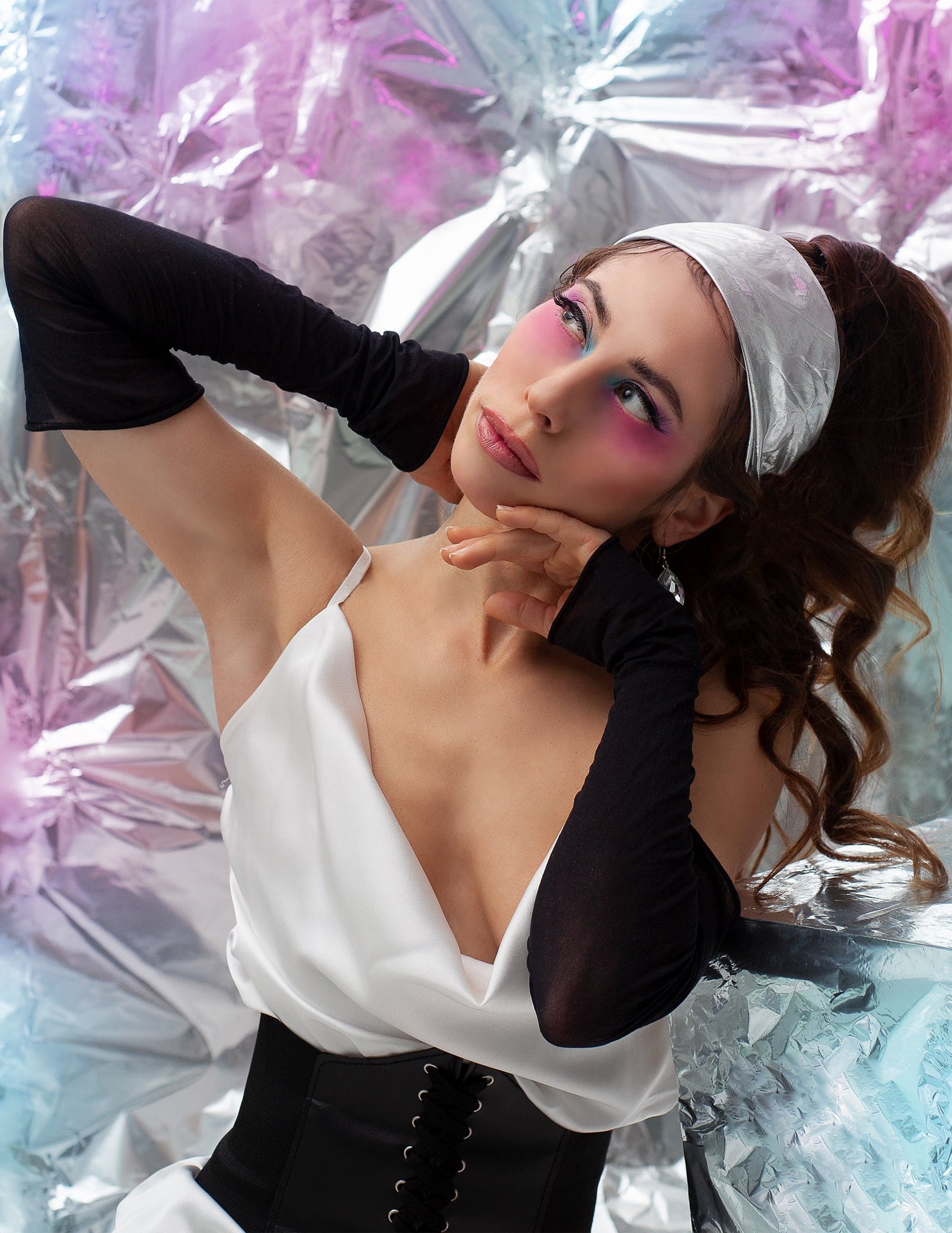 Ragazza con fascia argentata, trucco rosa e guanti neri, in top bianco e corsetto, in posa su un fondo di carta metallizzata iridescente