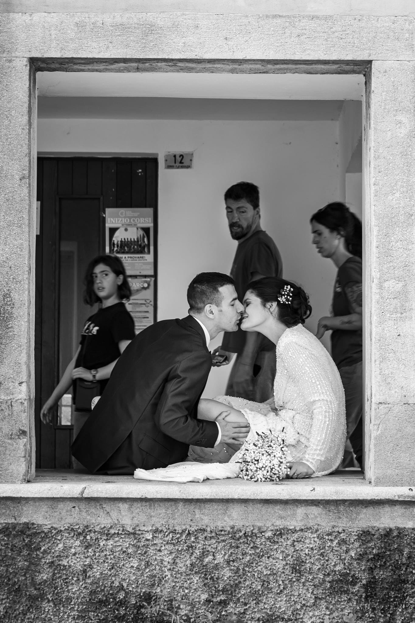 Sposi in bianco e nero si baciano seduti sul davanzale di una finestra, mentre due persone li osservano dall'interno