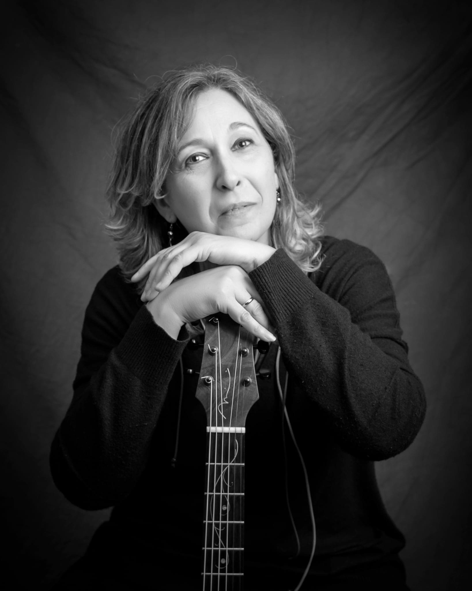In bianco e nero, donna dai capelli mossi con le mani incrociate sul manico di una chitarra, in maglione scuro su fondo sfumato