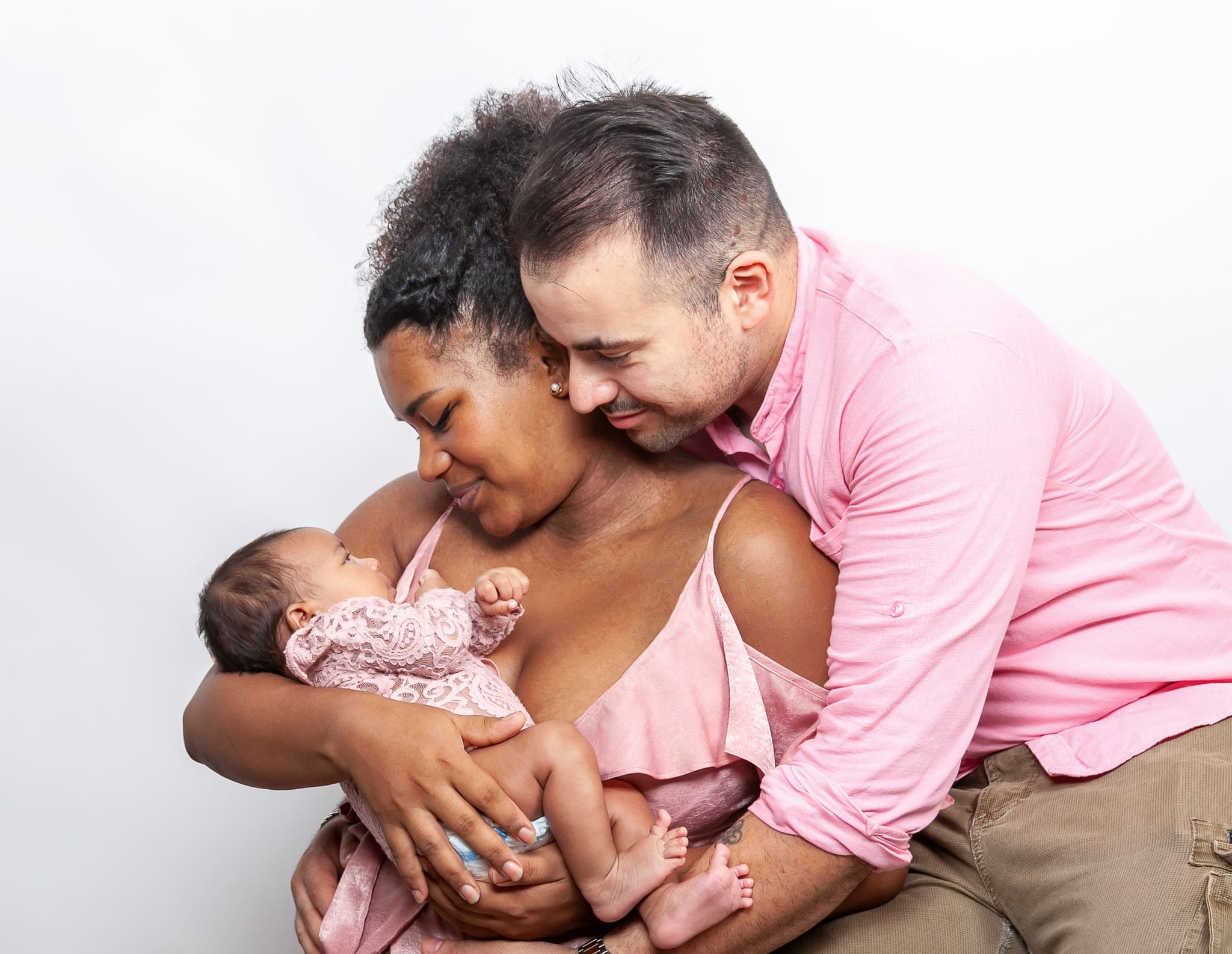 Mamma e papà abbracciano con dolcezza la loro neonata in pizzo rosa, su sfondo bianco, durante un servizio newborn in studio.