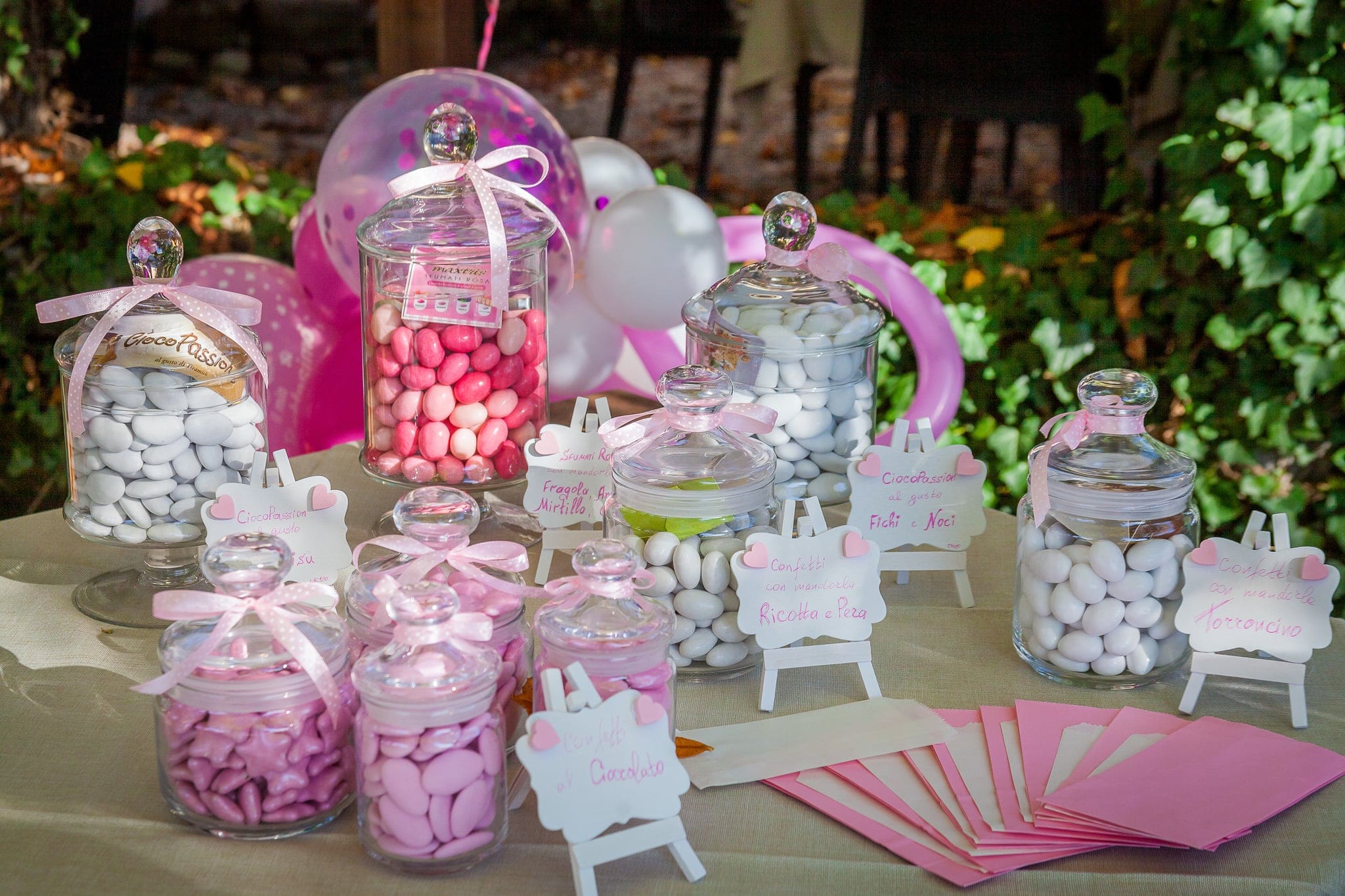 Tavolo della confettata con barattoli di vetro pieni di confetti rosa e bianchi, nastri e piccoli cartellini, tra i palloncini