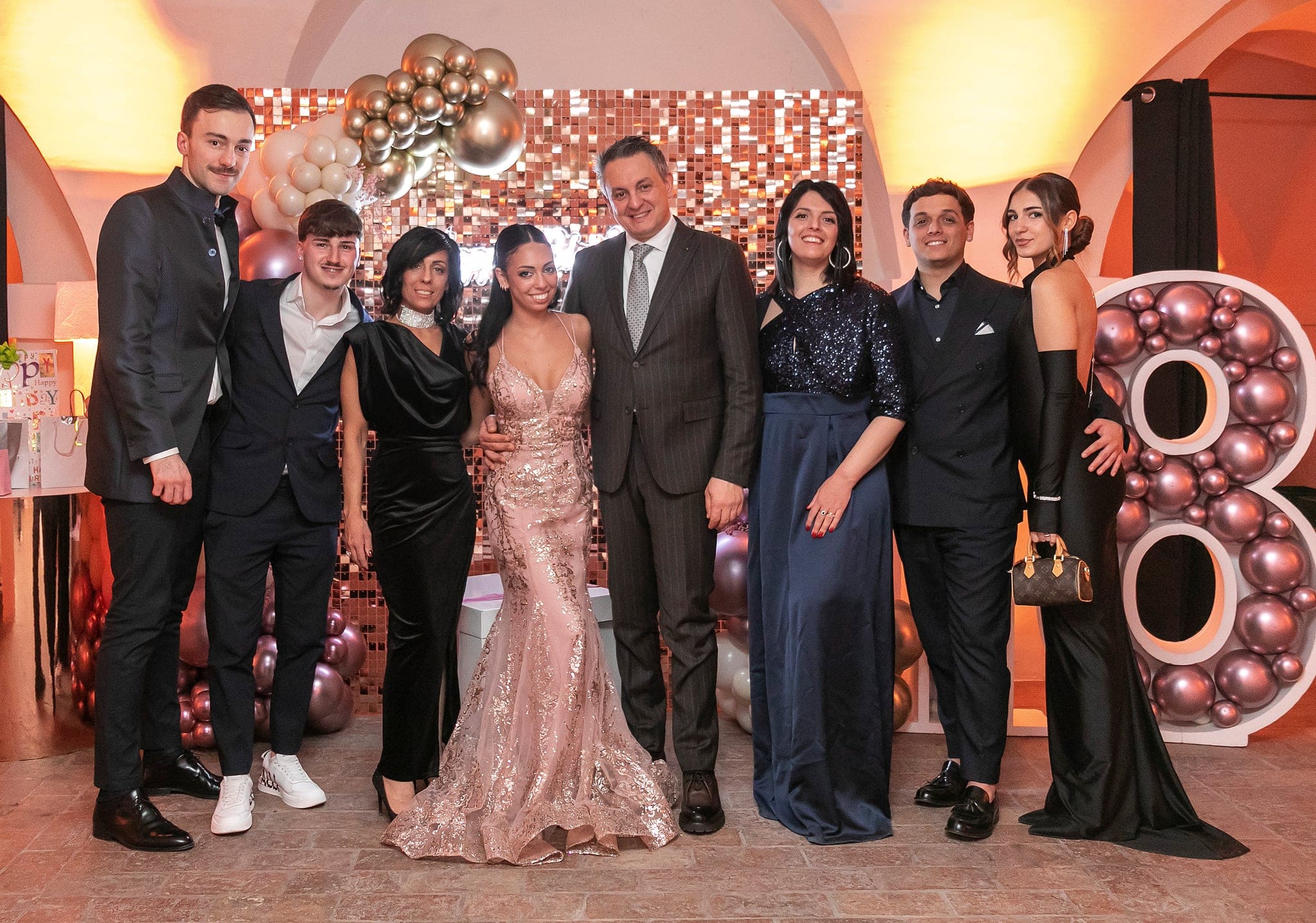 Foto di gruppo con la festeggiata al centro, familiari e amici in abiti eleganti davanti alla parete dorata e al numero 18