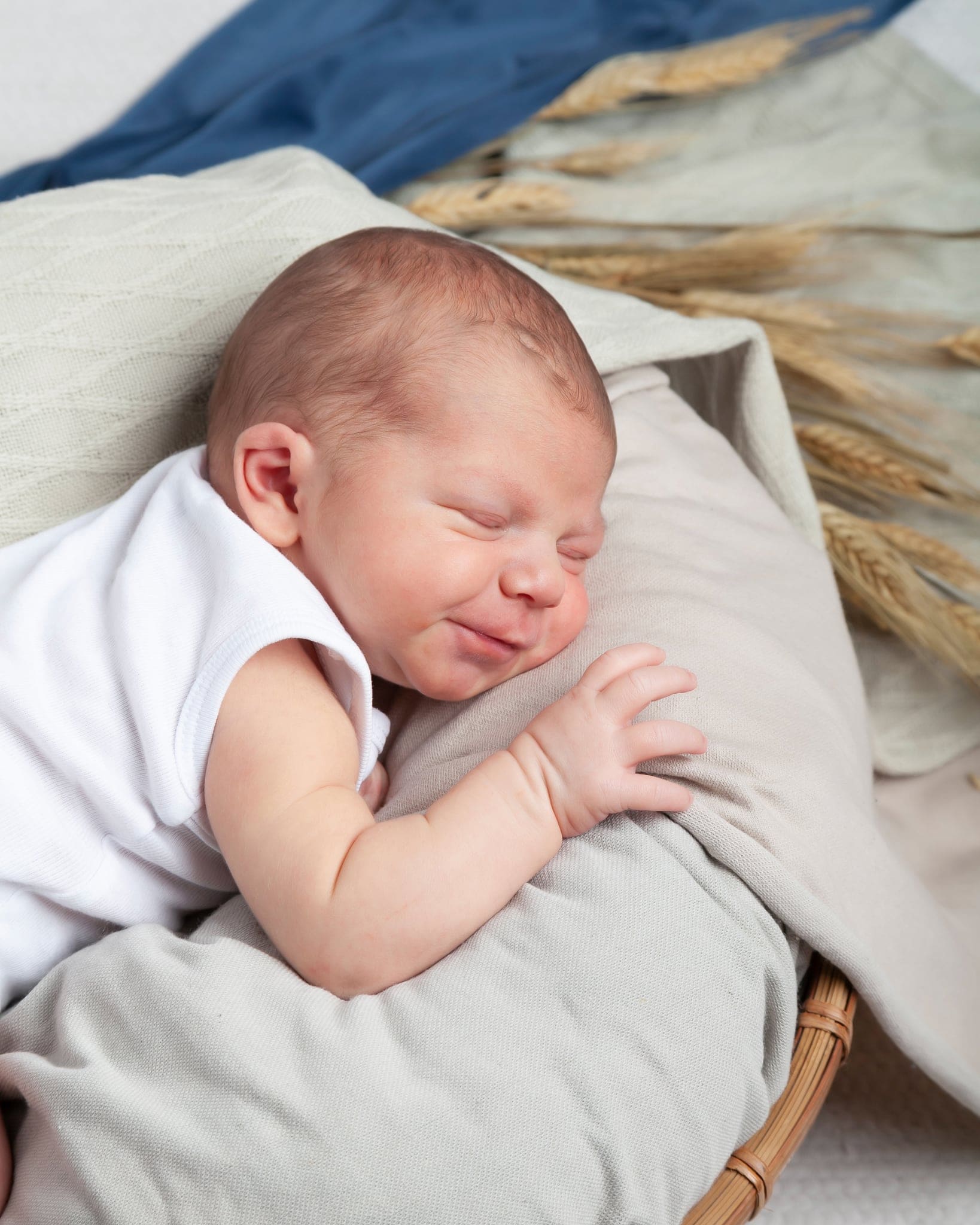 Neonato in body bianco che sorride nel sonno, sdraiato su un cuscino grigio con spighe di grano e un telo blu dietro.