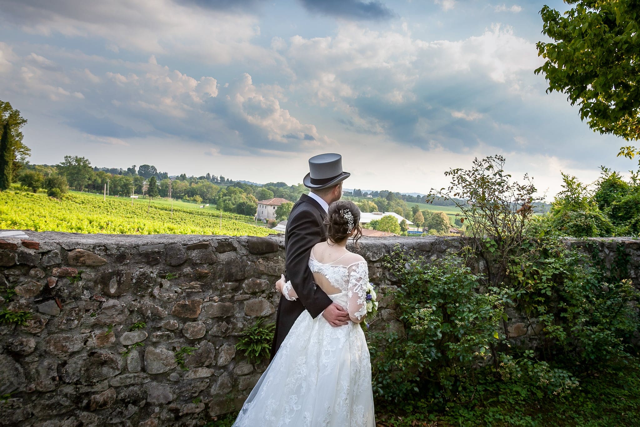 Sposi abbracciati di spalle davanti a un muretto di pietra con vista sui vigneti, lui con cilindro, a Villa Liruti
