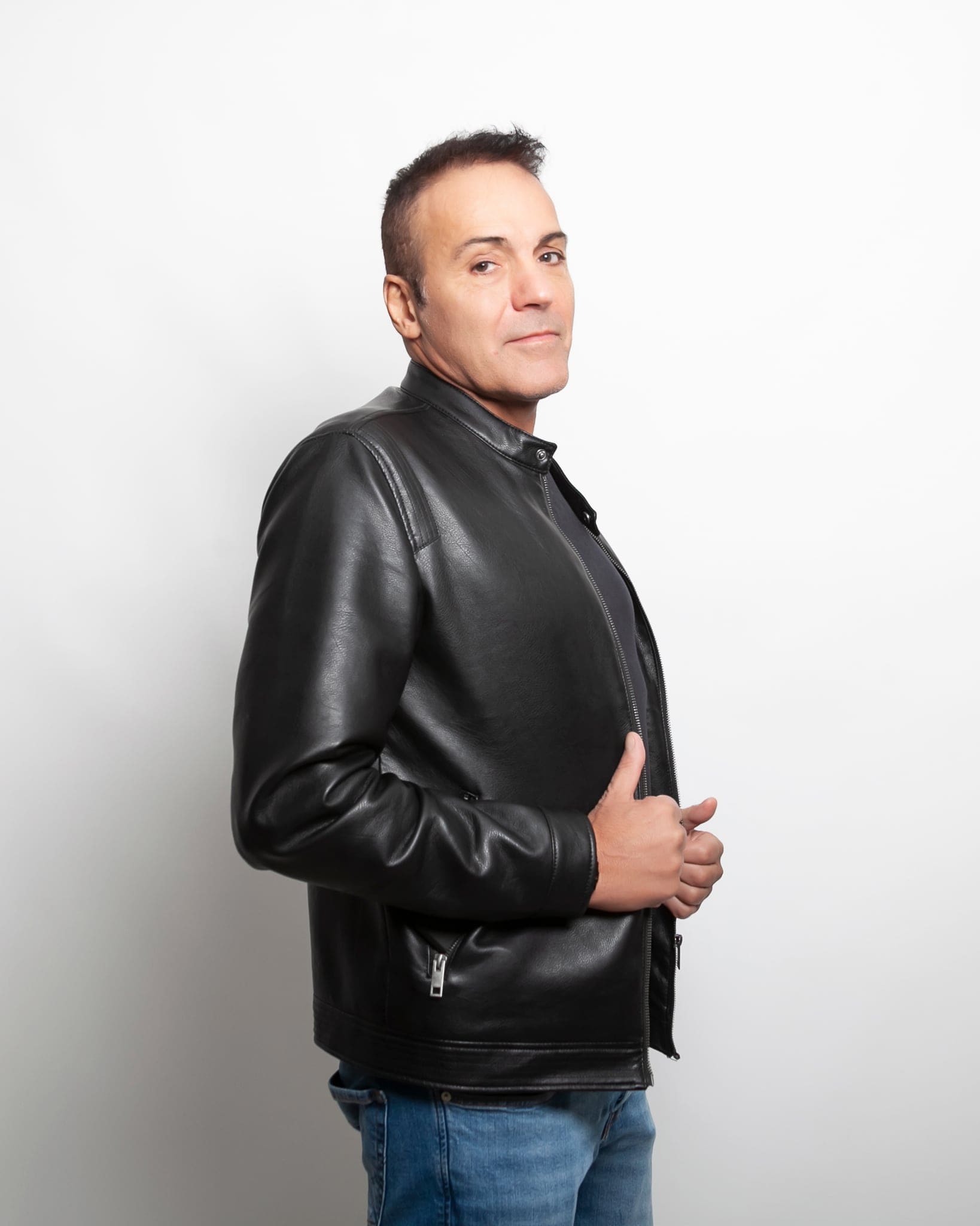 Uomo in giacca di pelle nera e jeans, di profilo con lo sguardo in camera, ritratto in studio su fondo bianco
