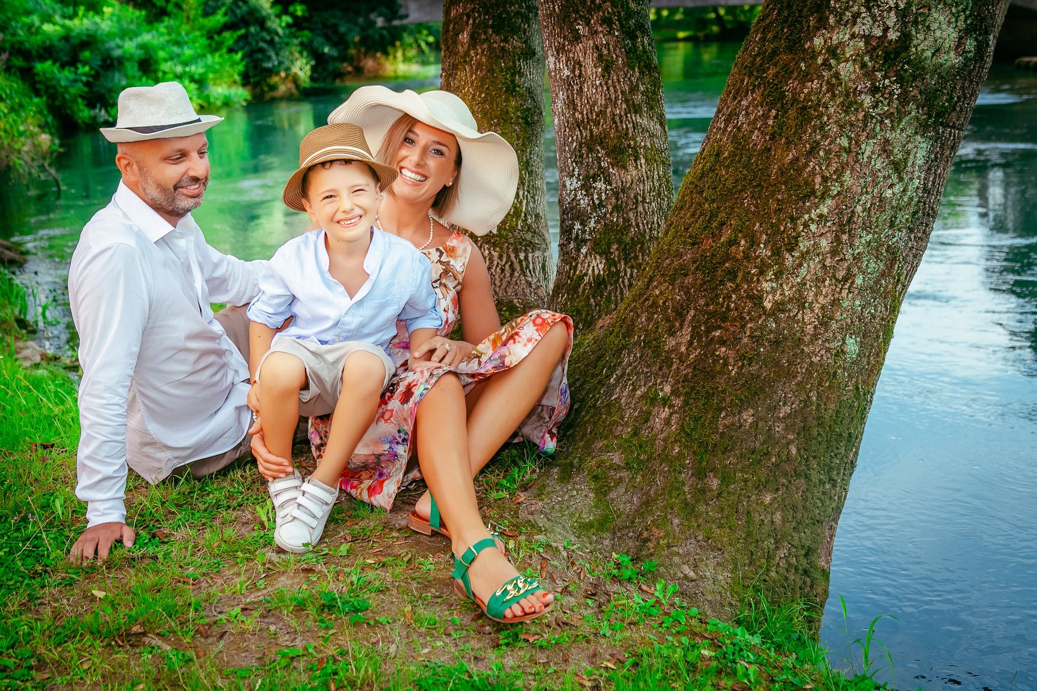 Famiglia con cappelli di paglia seduta sull'erba accanto a un albero sulla riva di un fiume turchese, bimbo sorridente