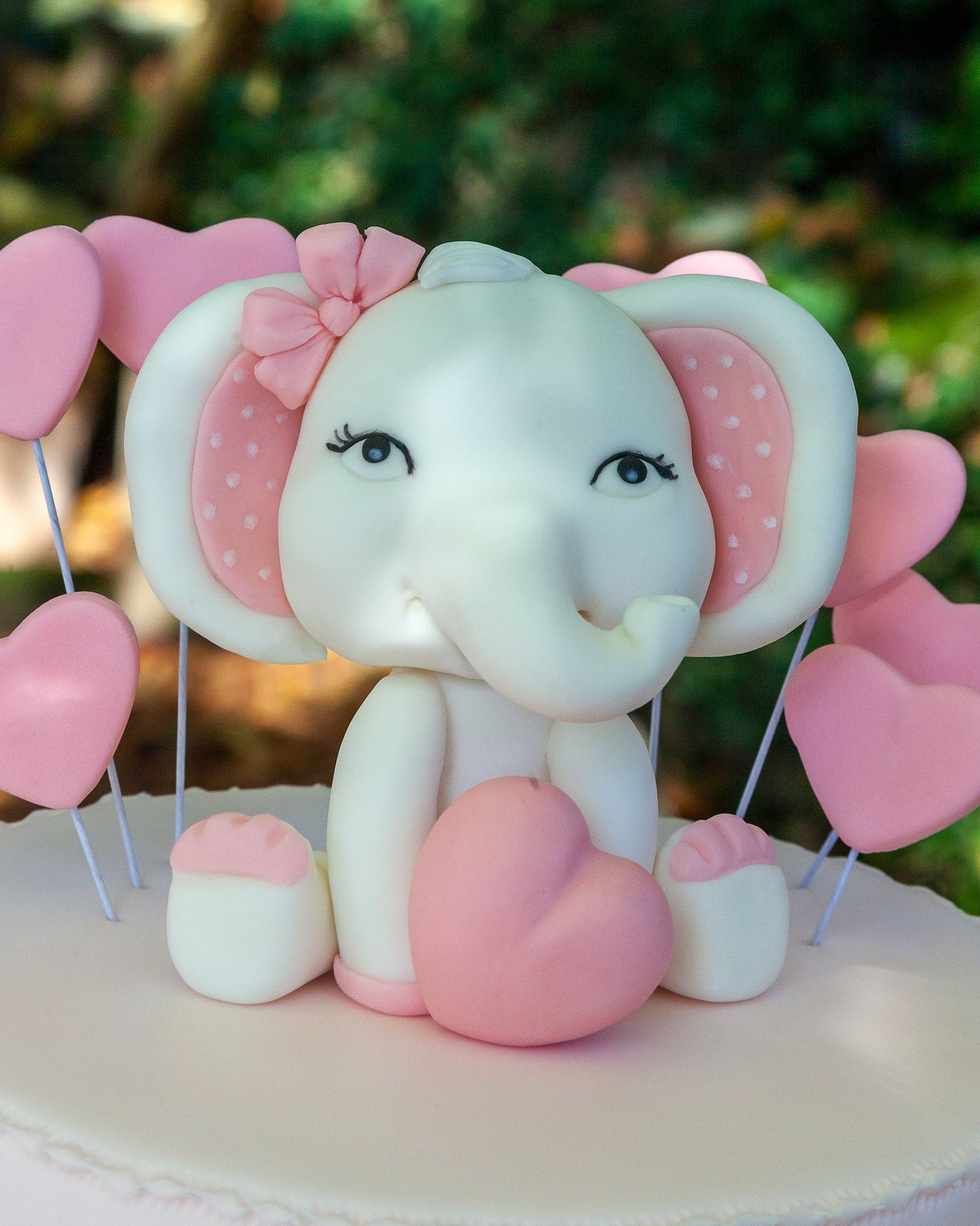 Primo piano di un elefantino in pasta di zucchero con fiocco e cuori rosa, sopra la torta del battesimo