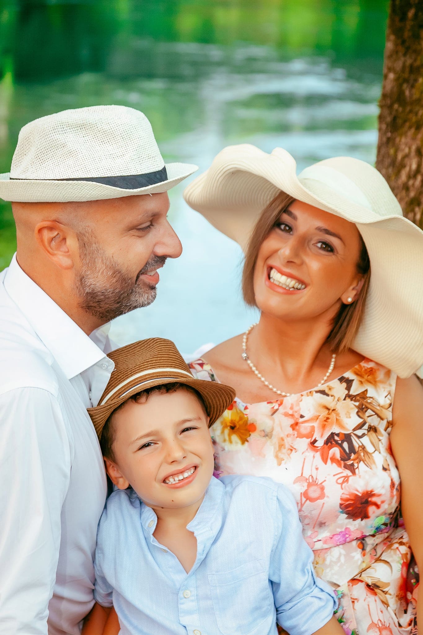 Ritratto ravvicinato di papà, mamma e figlio con cappelli estivi, sorridenti, davanti all'acqua verde di un fiume