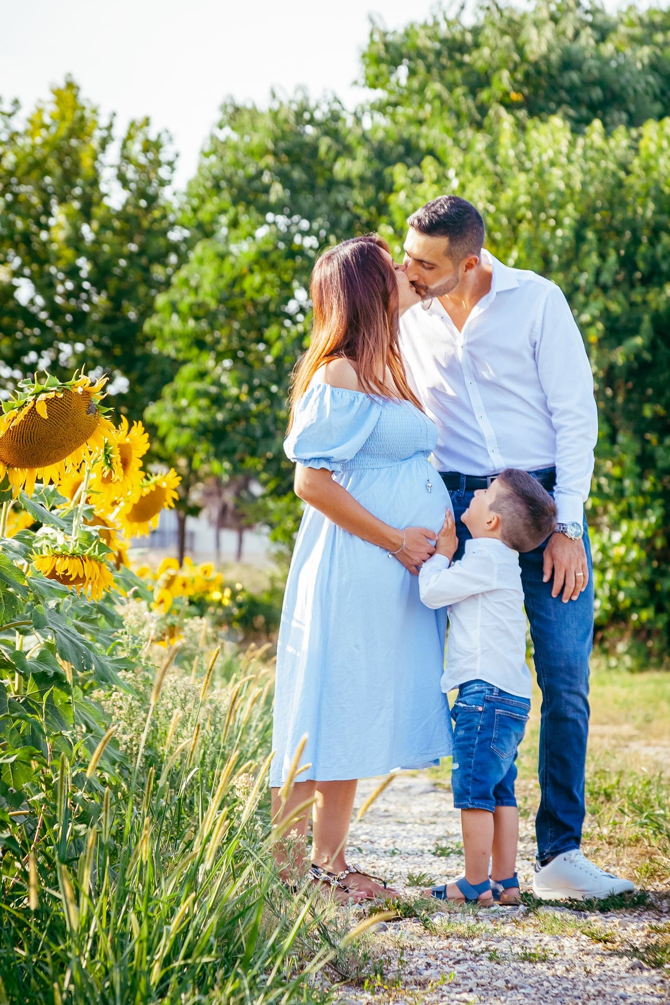 Futuri genitori che si baciano in un campo di girasoli con il figlio che li guarda, nella luce del tardo pomeriggio.