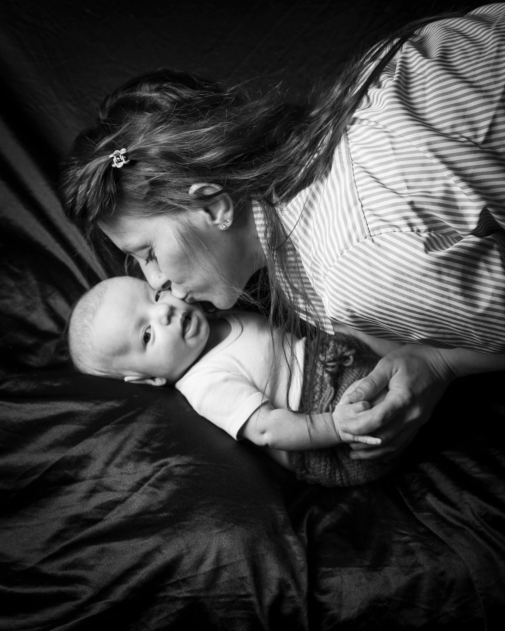 Mamma sdraiata che bacia sulla guancia il suo bebè con lo sguardo vivace, in bianco e nero su un tessuto scuro.