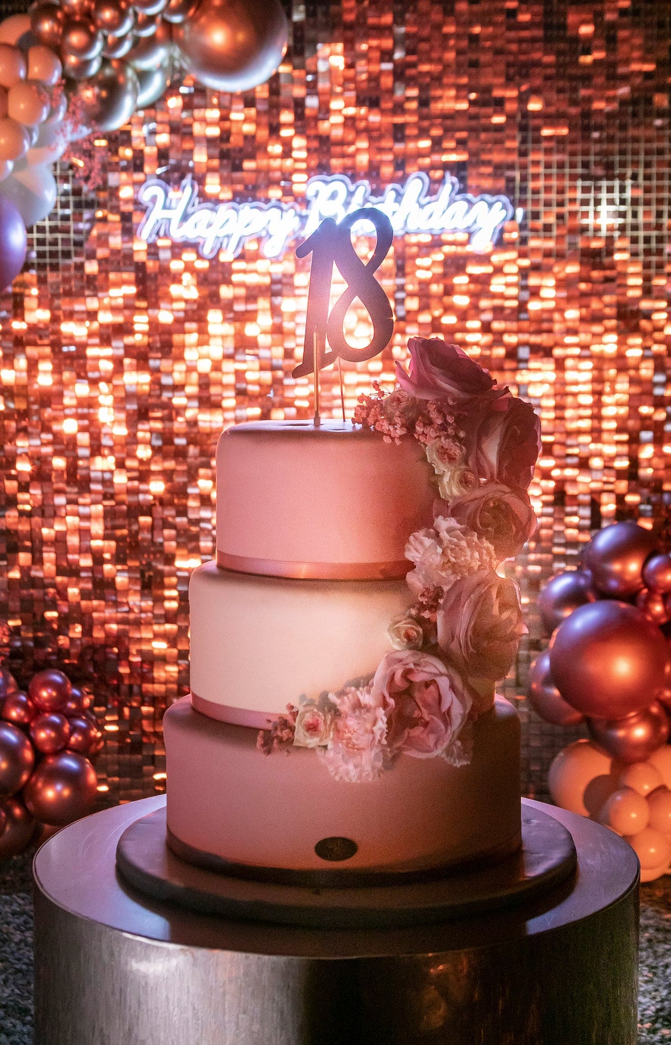 Torta a tre piani rosa e bianca decorata con rose e un 18 nero in cima, davanti alla parete di paillettes e ai palloncini