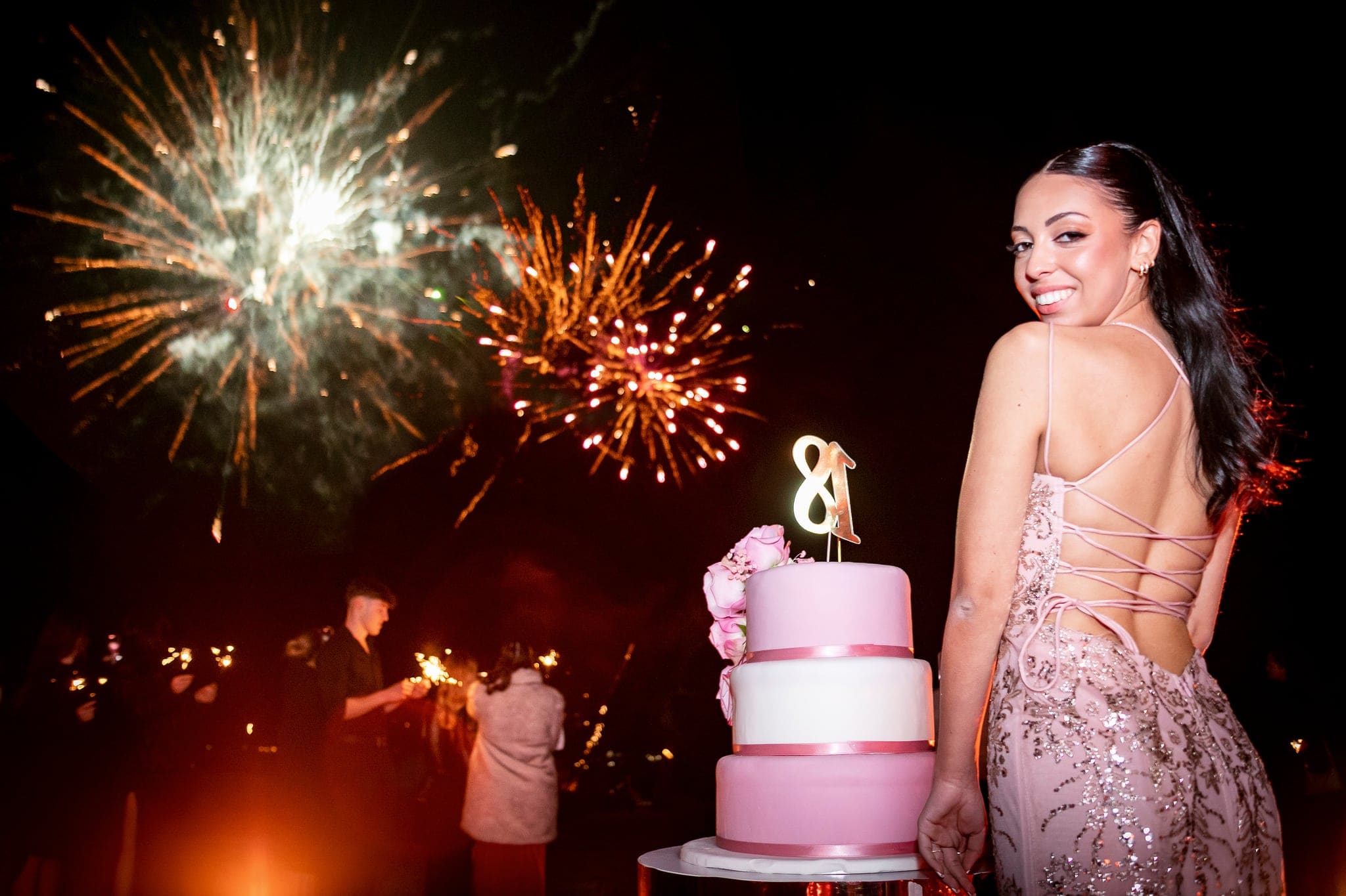 Ragazza in abito rosa con schiena in pizzo si volta sorridendo accanto alla torta, mentre i fuochi d'artificio illuminano il cielo notturno
