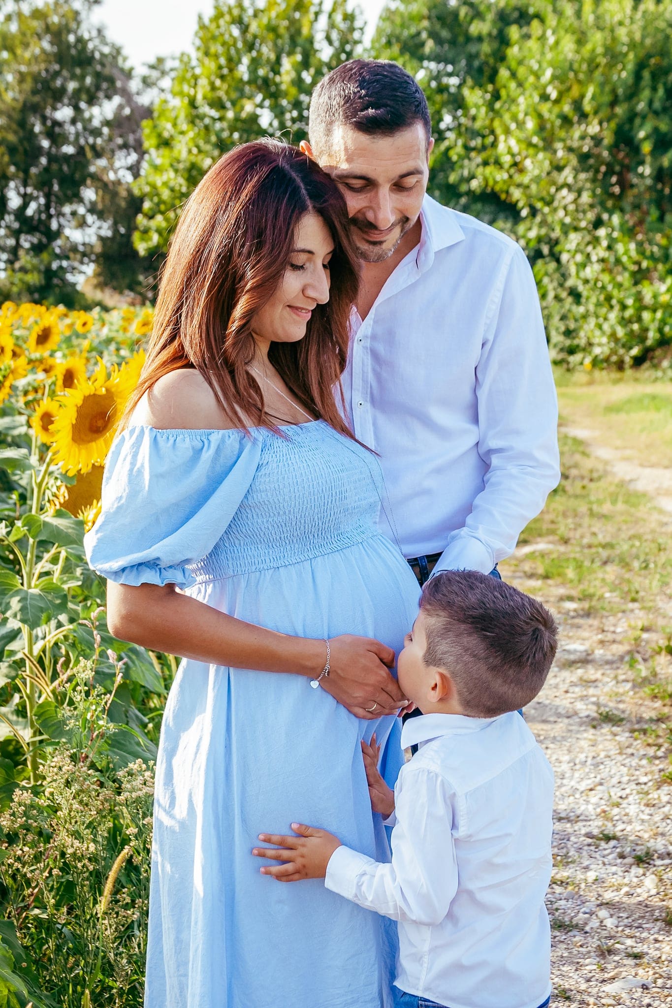 Mamma in abito azzurro con papà e figlioletto che le bacia il pancione, tra i girasoli lungo un sentiero di campagna.