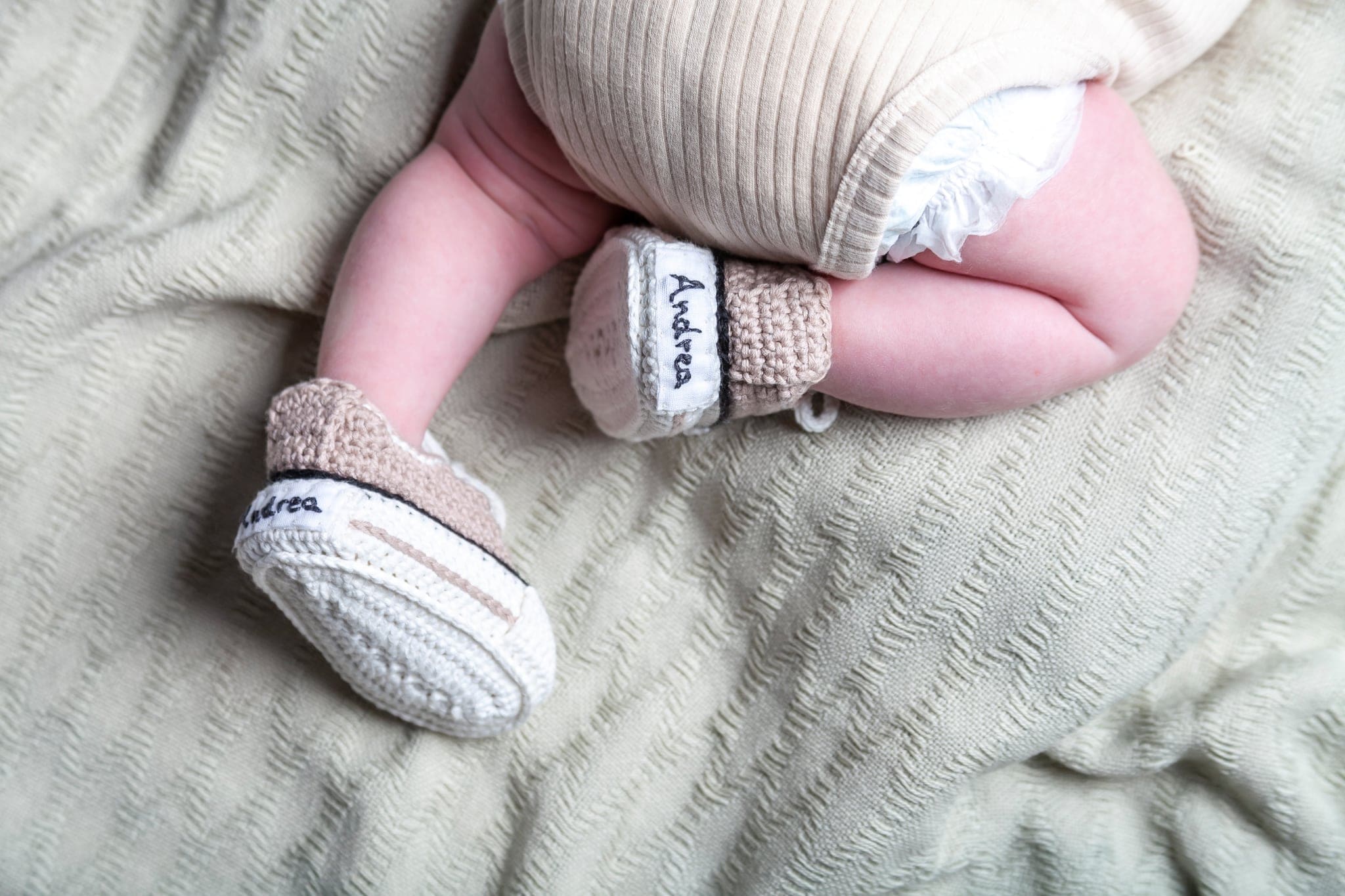 Gambe di un neonato con scarpine di lana in stile sneaker con un nome ricamato, su un morbido plaid verde chiaro.