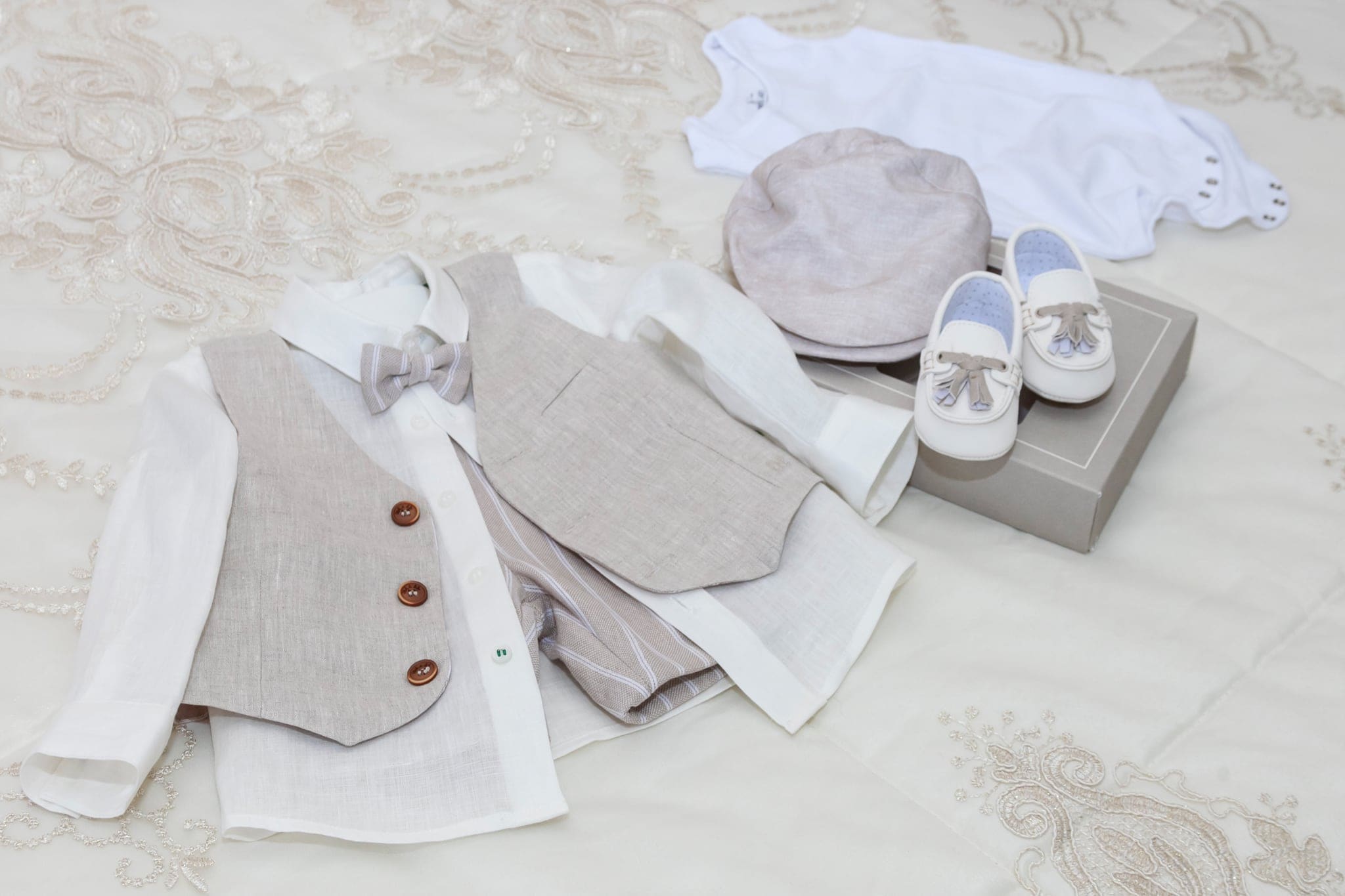 Camicia, gilet beige, cappellino e scarpine bianche del piccolo appoggiati sul copriletto ricamato, pronti per il battesimo