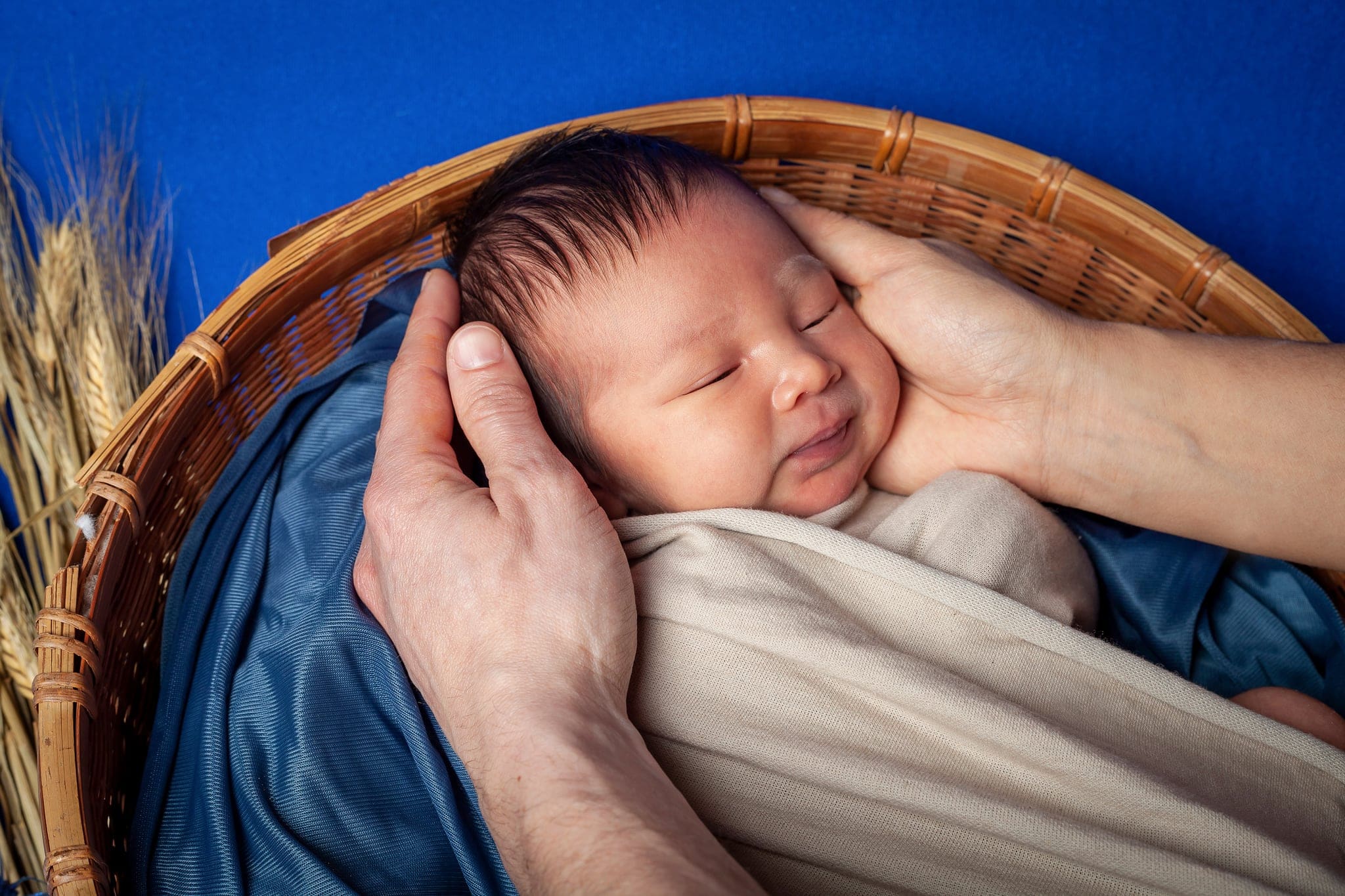 Neonato che dorme in un cesto con telo blu, il viso sostenuto delicatamente dalle mani del papà, con spighe sul fondo.