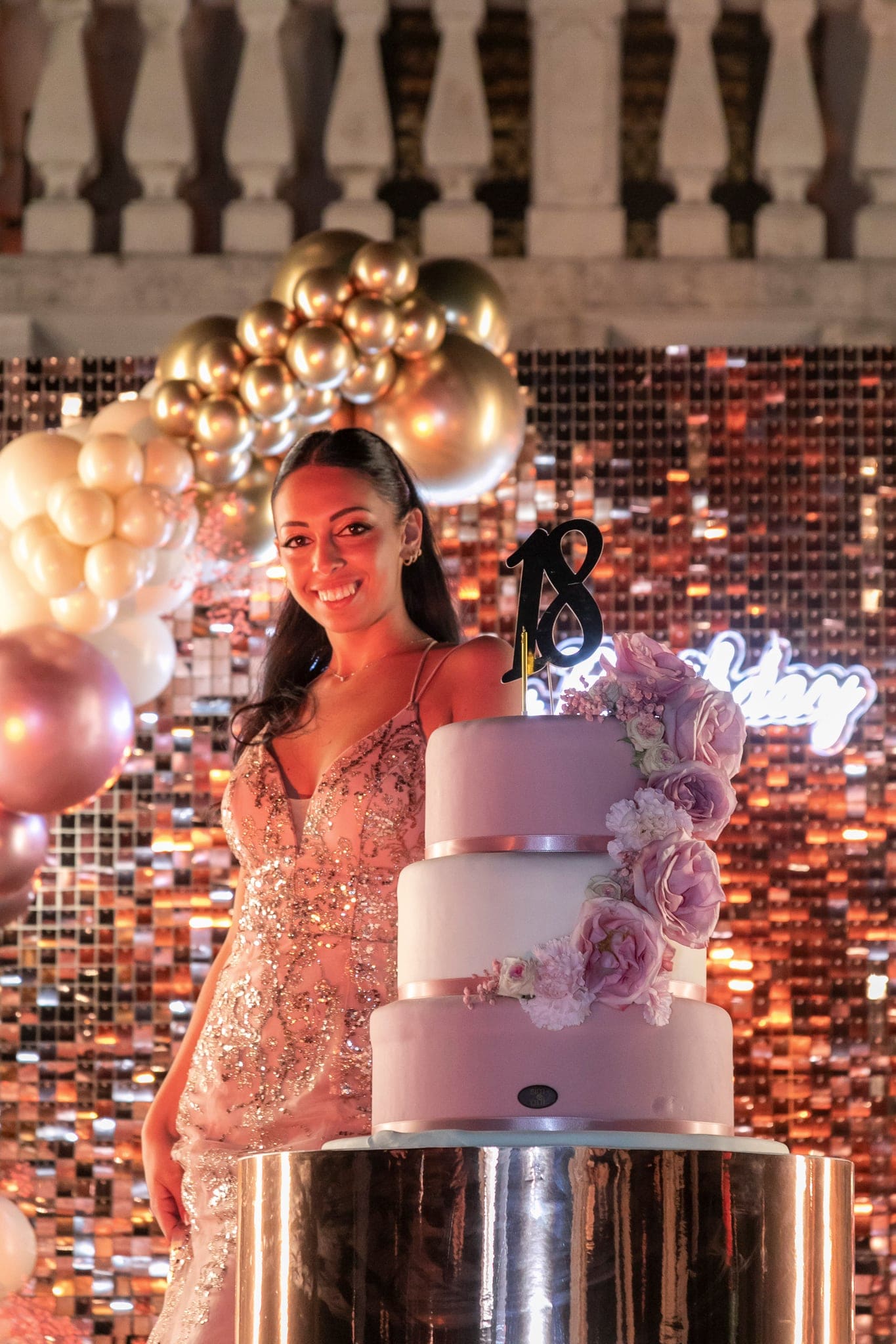 Ragazza sorridente in abito rosa dietro la torta con il 18 nero, davanti alla parete di paillettes e ai palloncini dorati