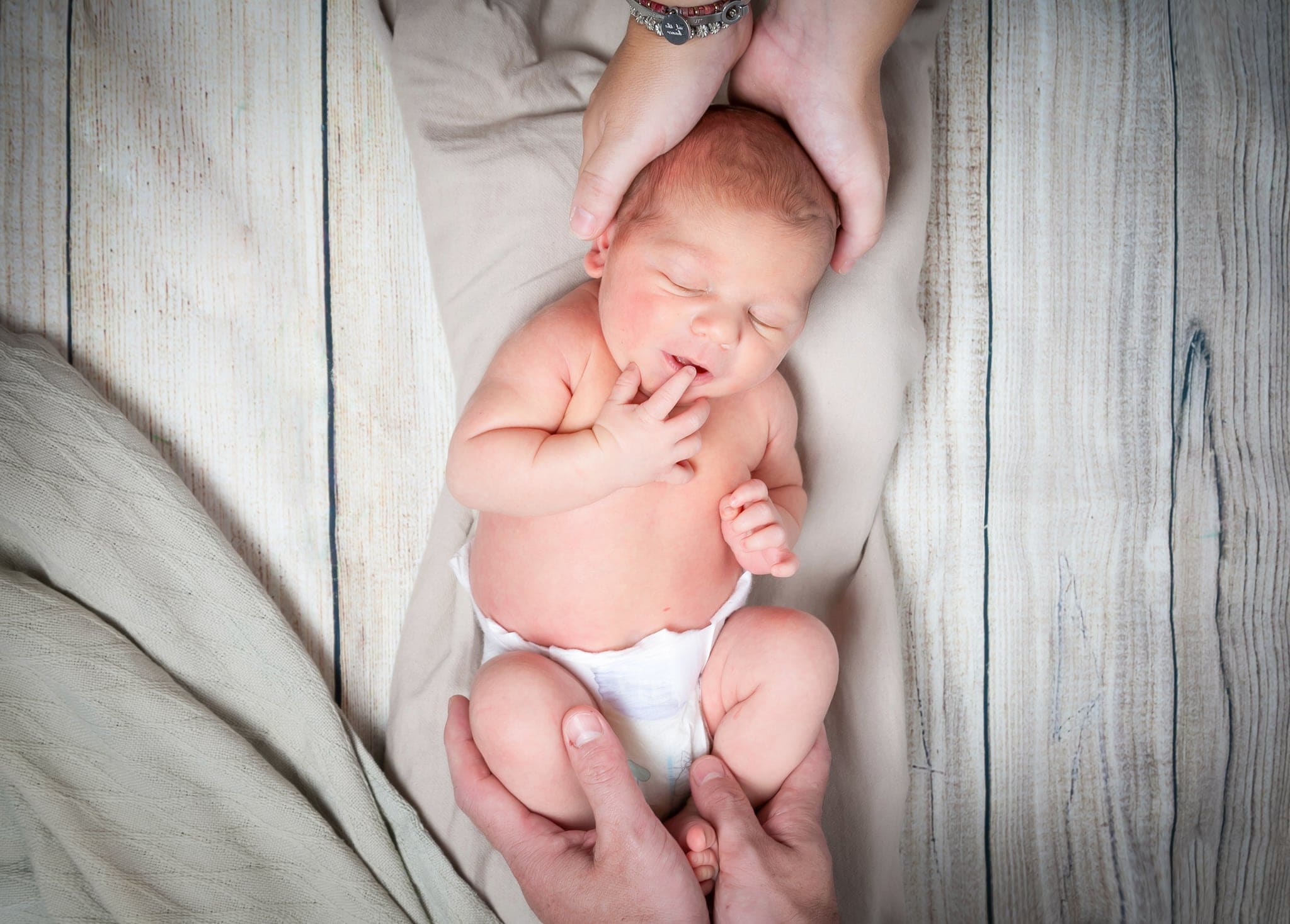 Neonato in pannolino che si succhia il dito su assi di legno chiaro, con le mani di un adulto che gli sostengono testa e gambe.