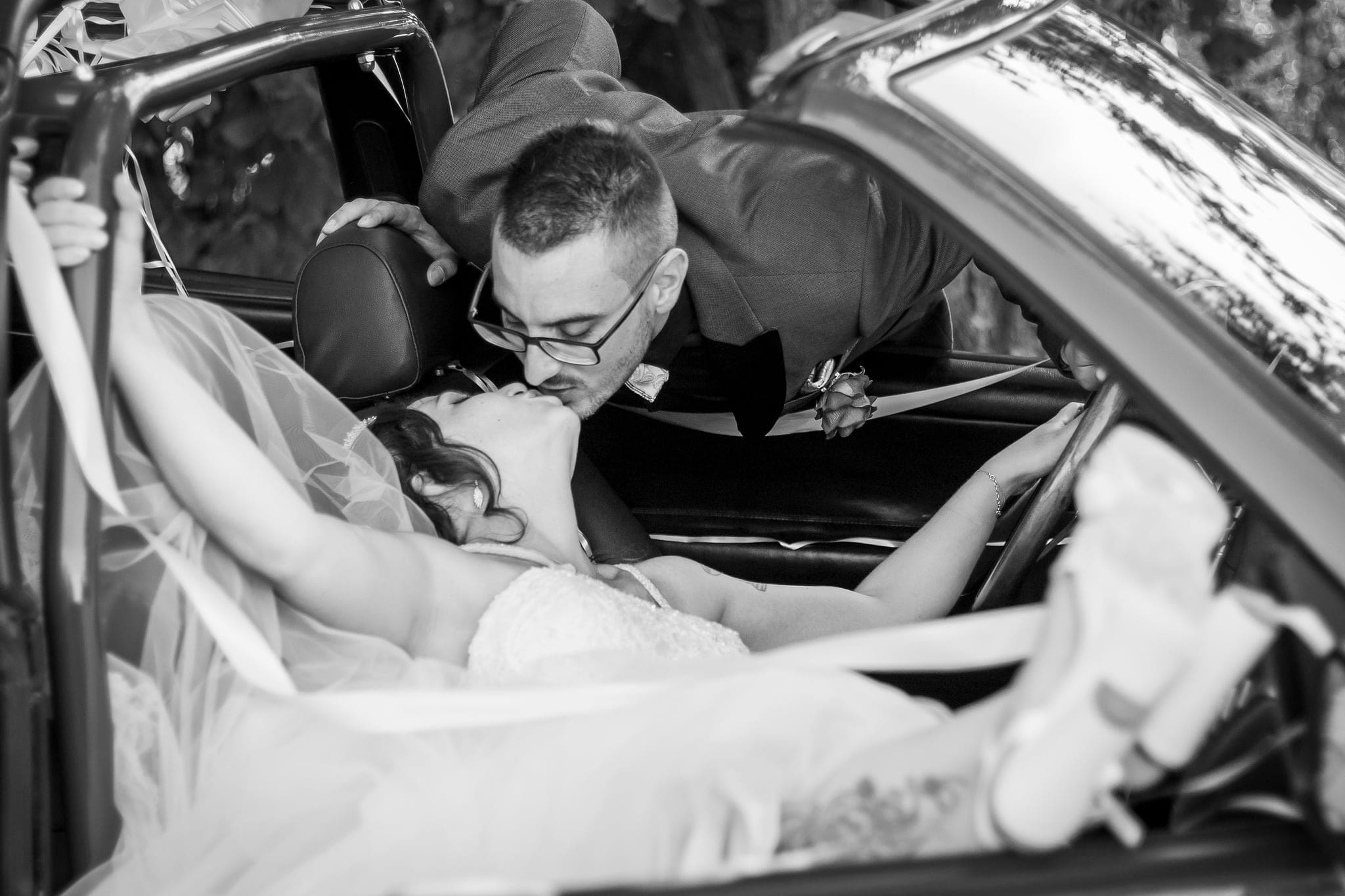 Sposo con occhiali bacia la sposa sdraiata sul sedile di un'auto d'epoca, in bianco e nero, con il velo mosso