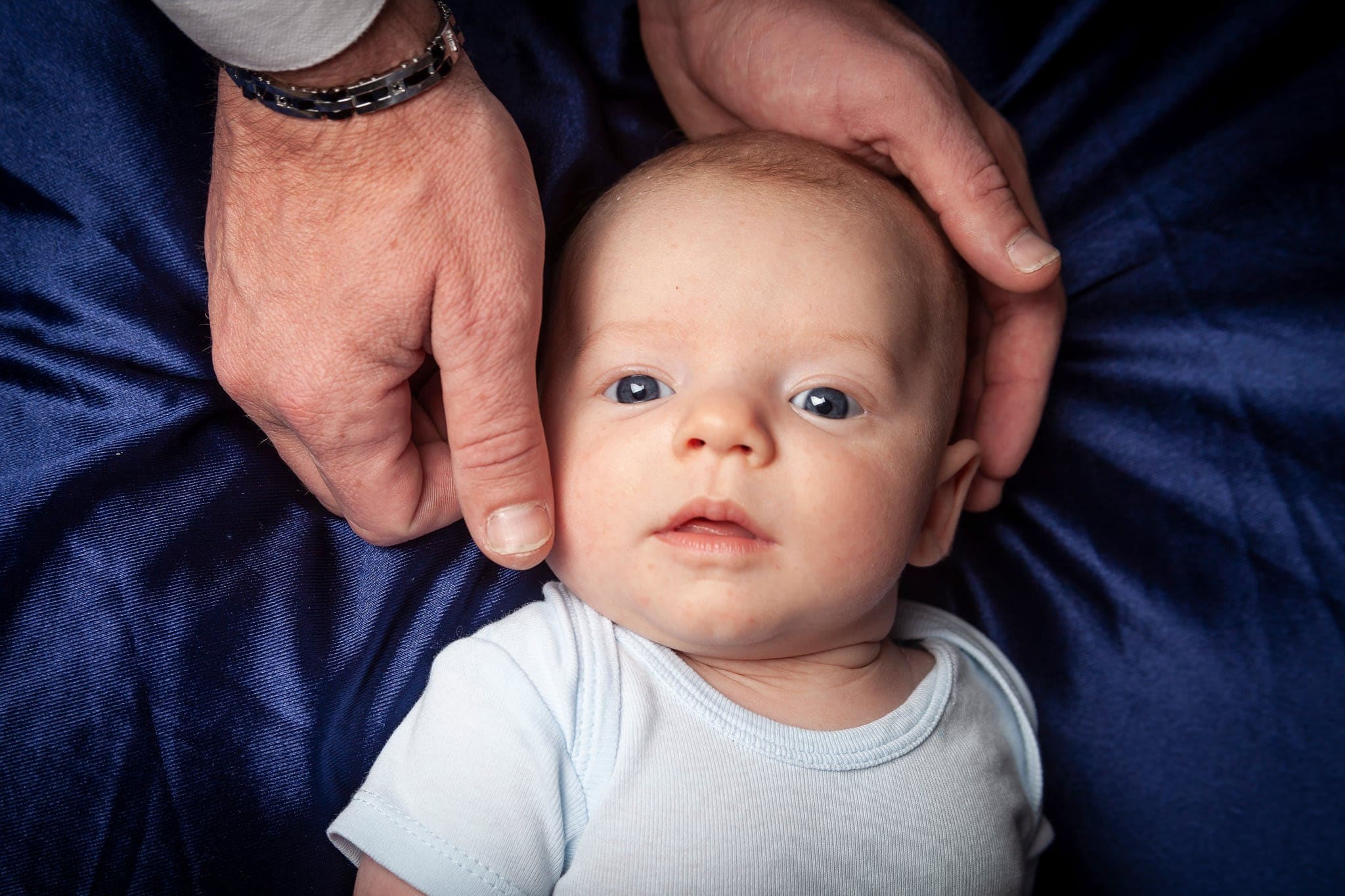 Primo piano di un bebè con occhi chiari in body azzurro, con le grandi mani del papà che gli sfiorano il viso, su raso blu.