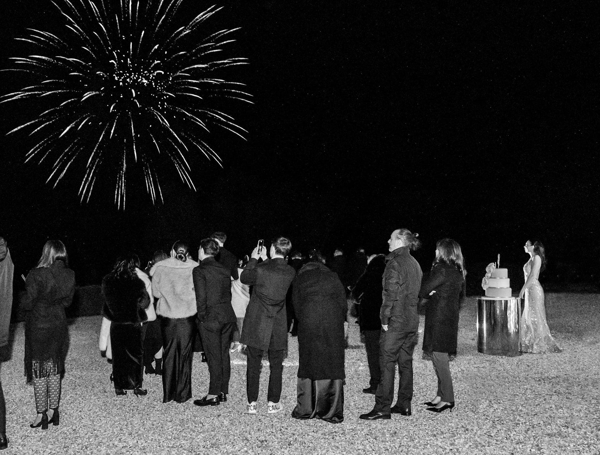 In bianco e nero, gli invitati e la festeggiata accanto alla torta guardano un grande fuoco d'artificio nel cielo scuro
