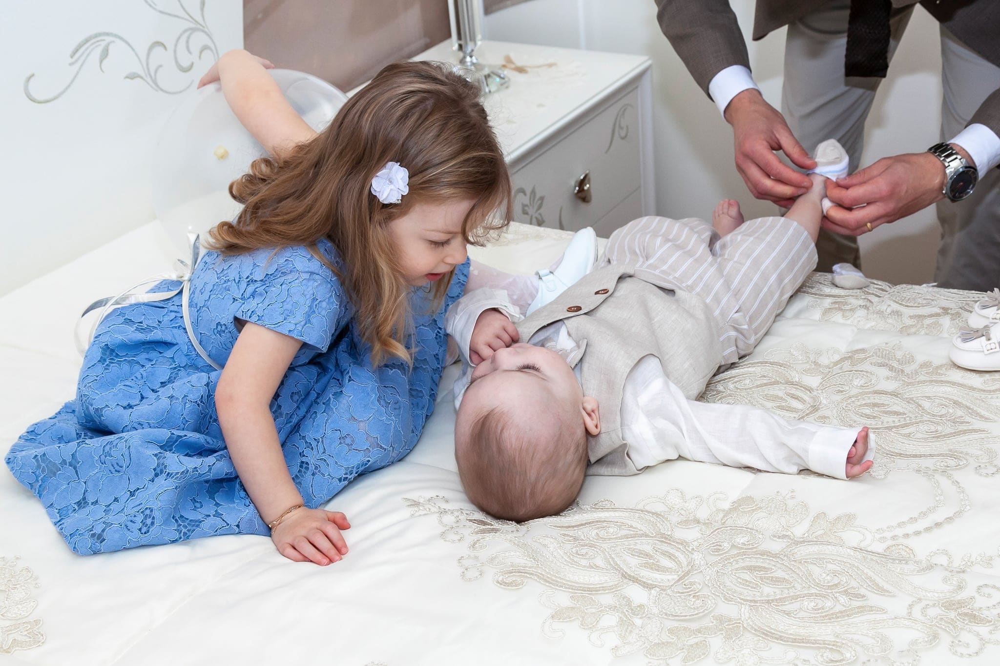 La sorellina in abito azzurro con un fiocco tra i capelli si china sul fratellino sdraiato sul letto mentre il papà lo veste