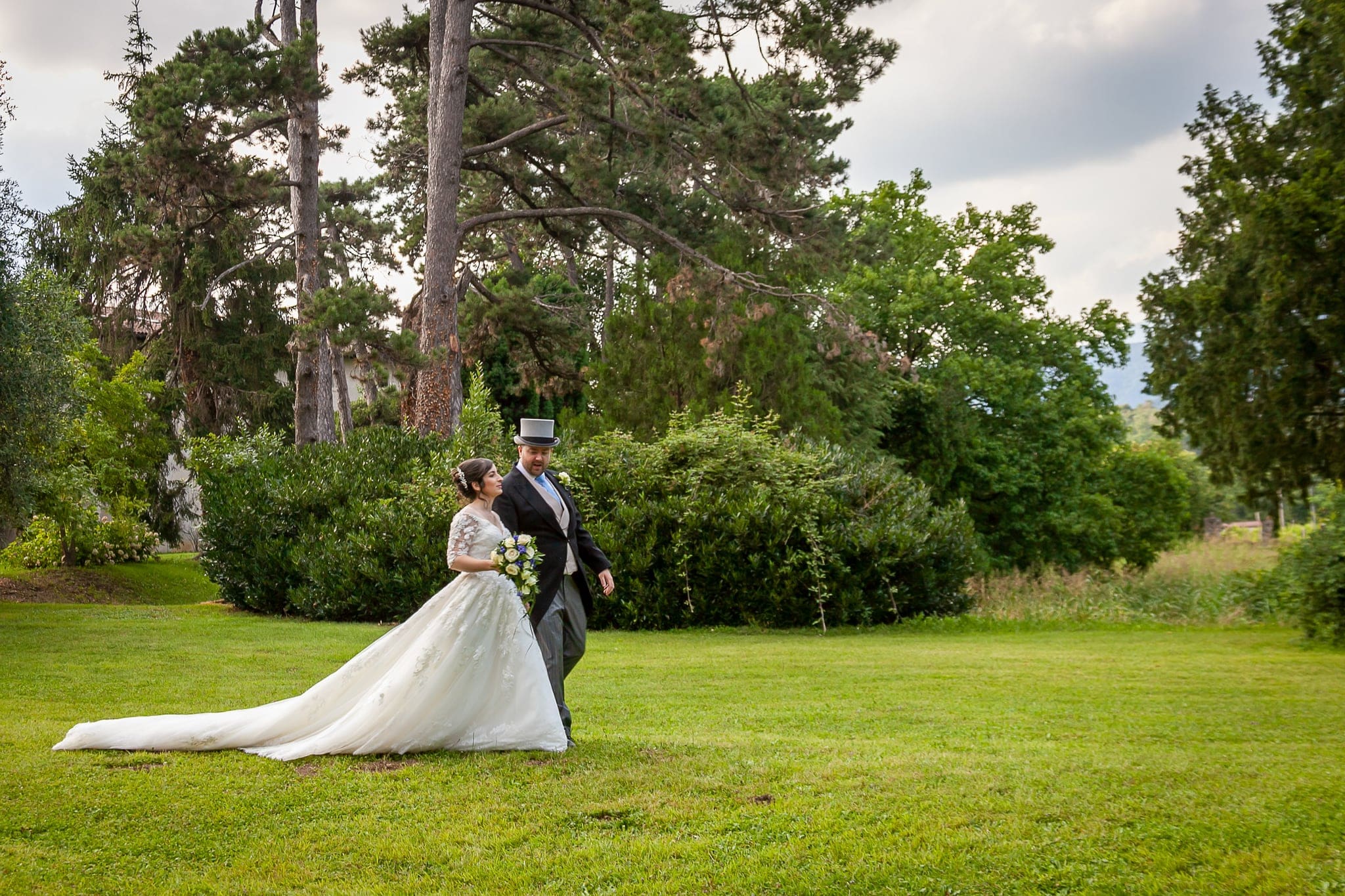 Sposa con lungo strascico e sposo con cilindro camminano su un prato all'ombra di grandi alberi, matrimonio a Corno di Rosazzo