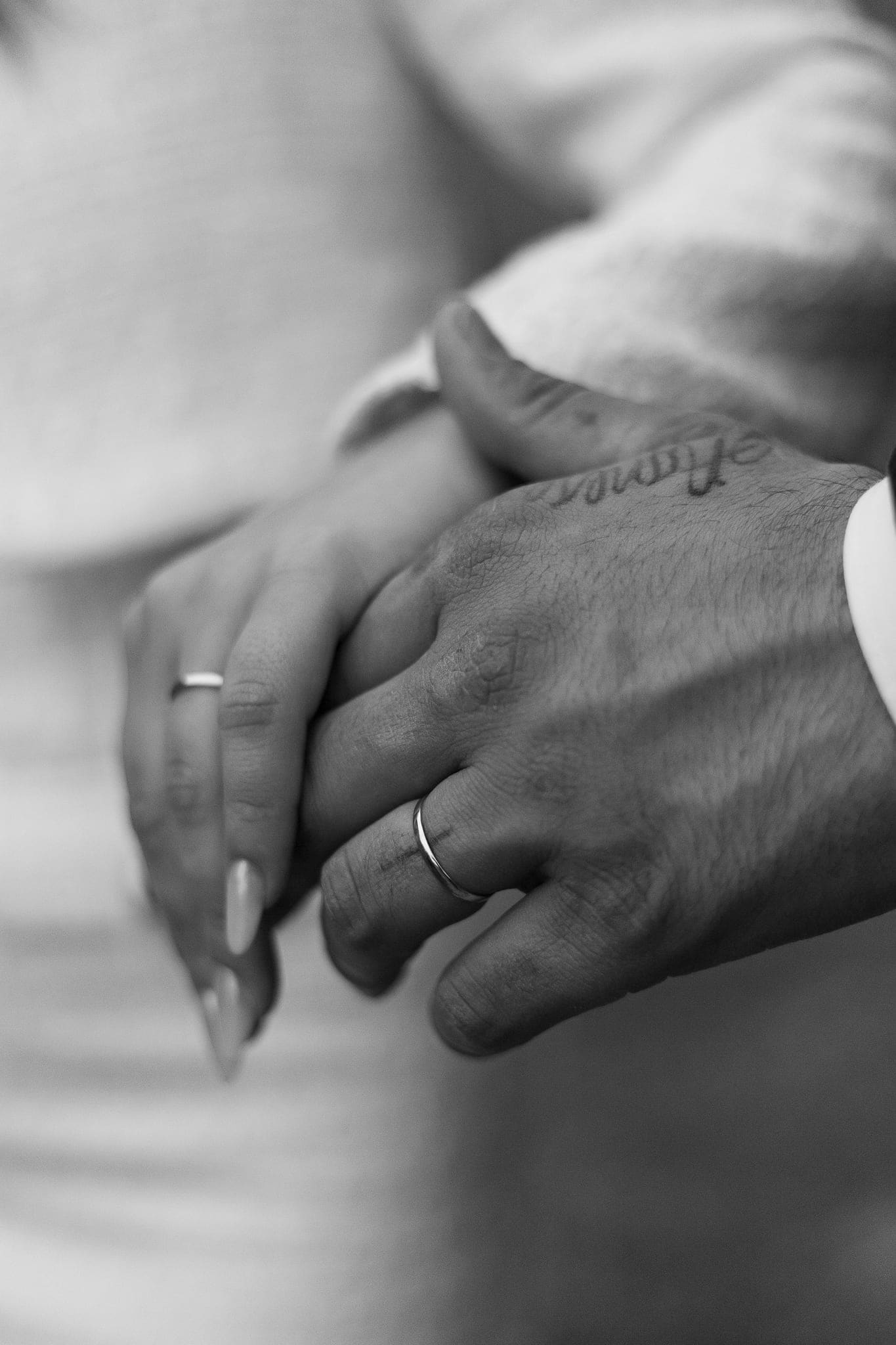 Primo piano in bianco e nero delle mani intrecciate degli sposi con le fedi nuziali, una con un tatuaggio sul dorso