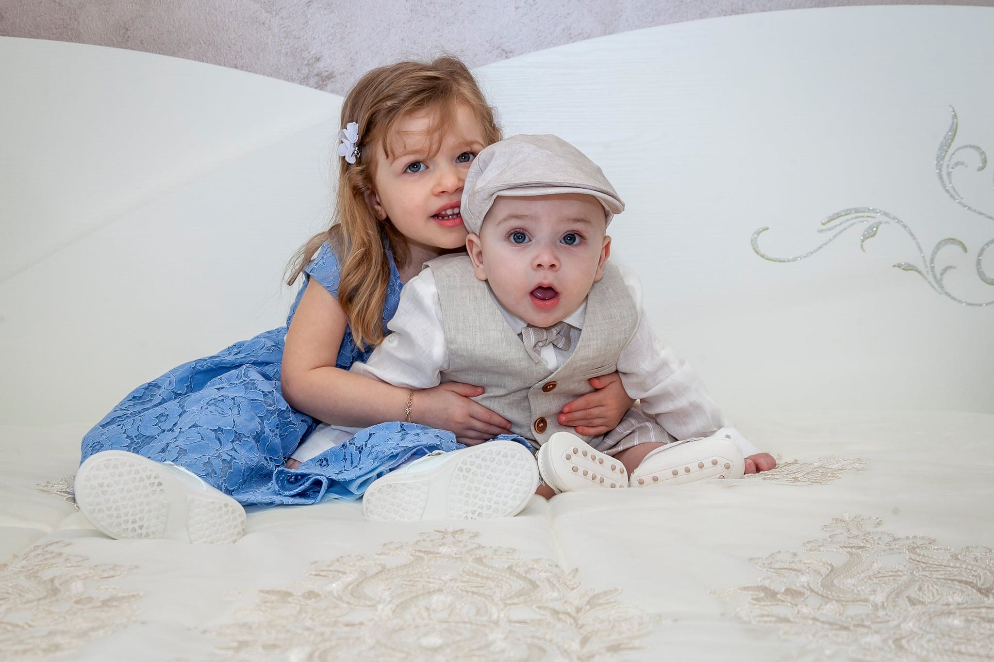 Seduti sul letto bianco, la sorellina in abito azzurro abbraccia il fratellino con cappellino e gilet, che guarda stupito