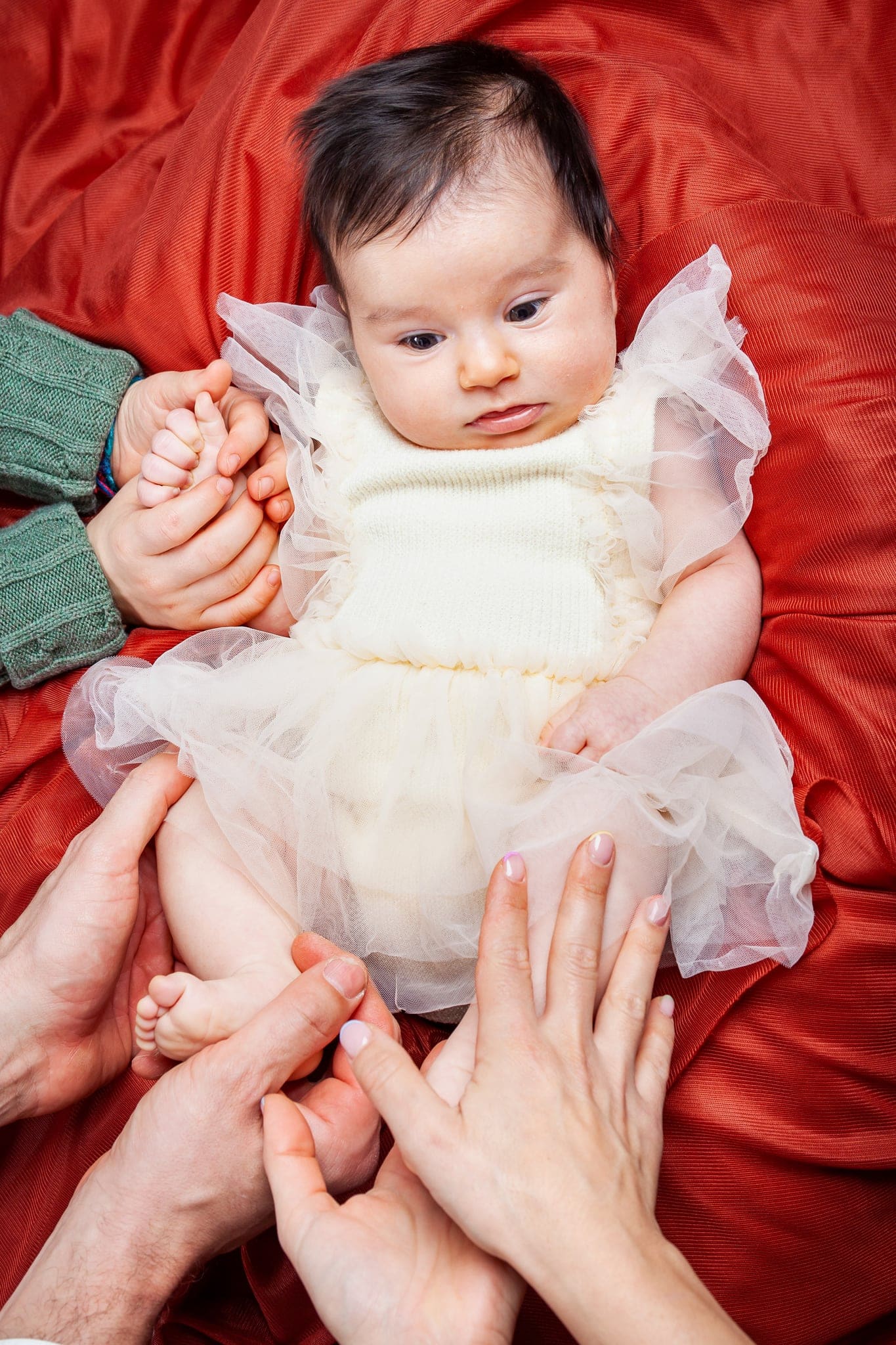 Bebè con abitino panna in tulle su telo rosso, con le manine di mamma e fratellino che le tengono mani e piedini.