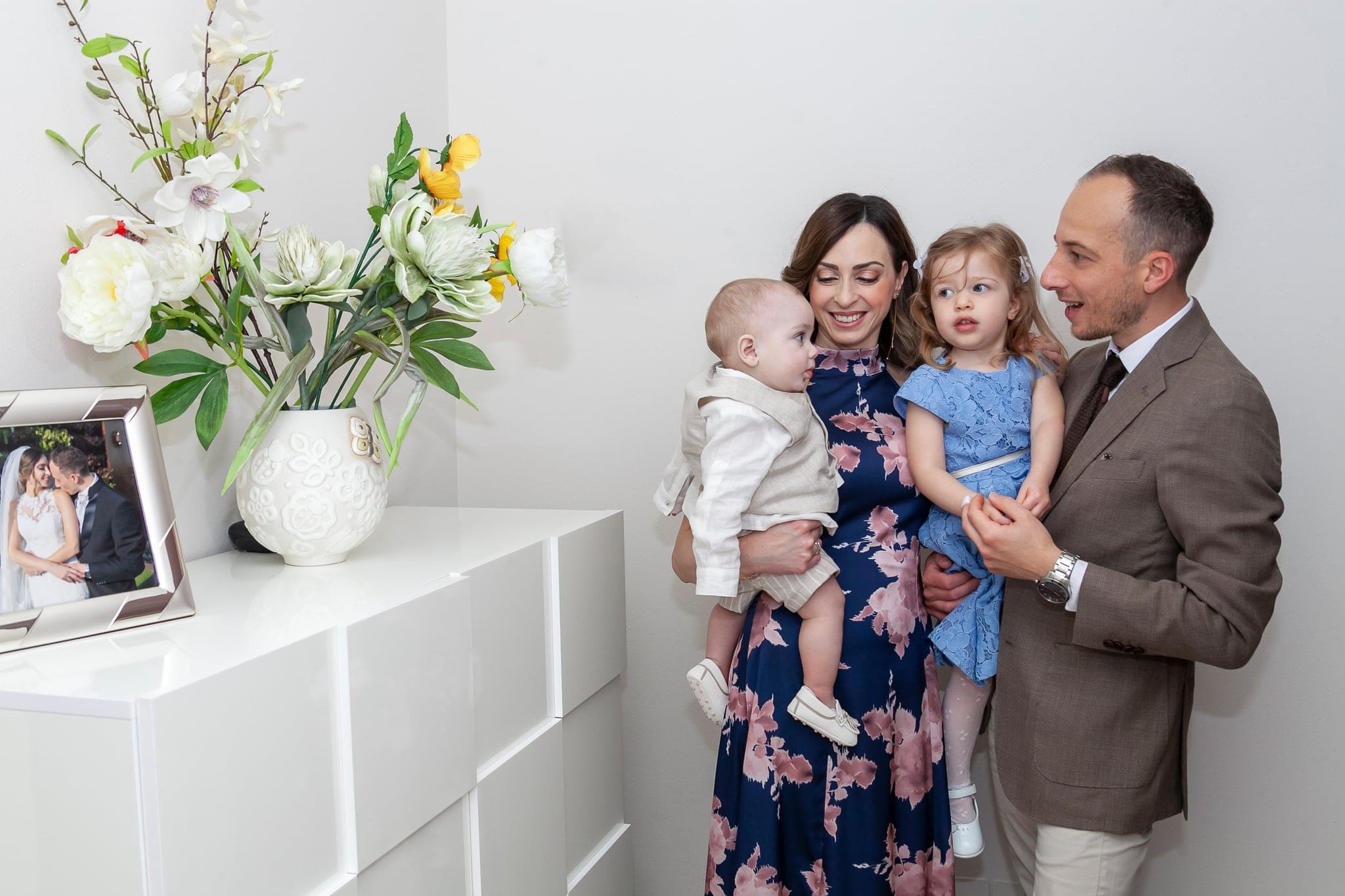Ritratto in casa di mamma, papà e due figli, il piccolo in braccio alla mamma, accanto a un vaso di fiori bianchi