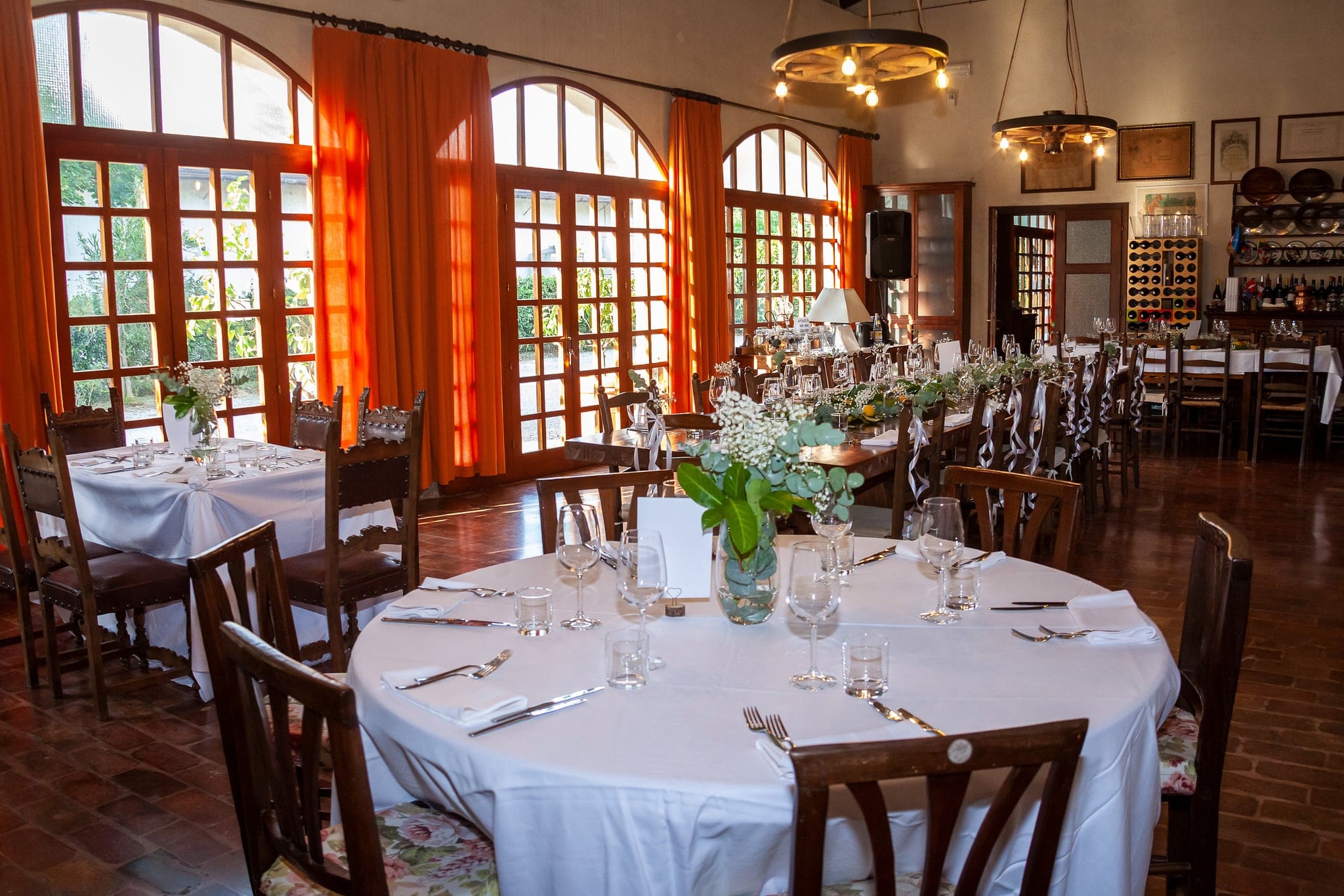 Sala di un ristorante con tavoli rotondi apparecchiati, tende arancioni e grandi finestre ad arco, matrimonio sul Collio Friulano