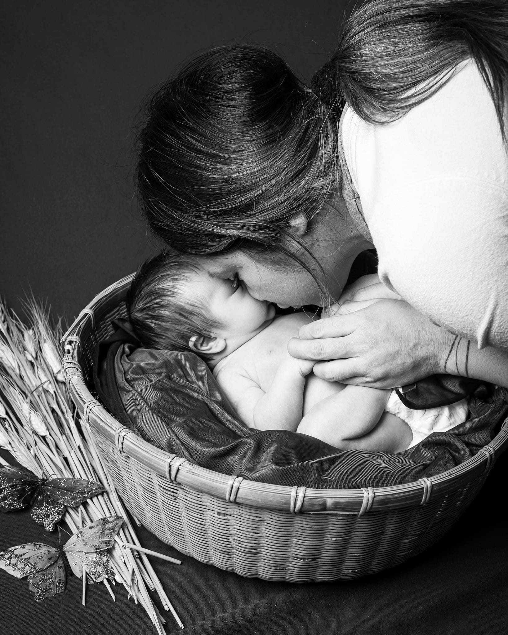 Mamma che si china a baciare il neonato in un cesto con spighe e farfalle, in bianco e nero su fondo scuro.