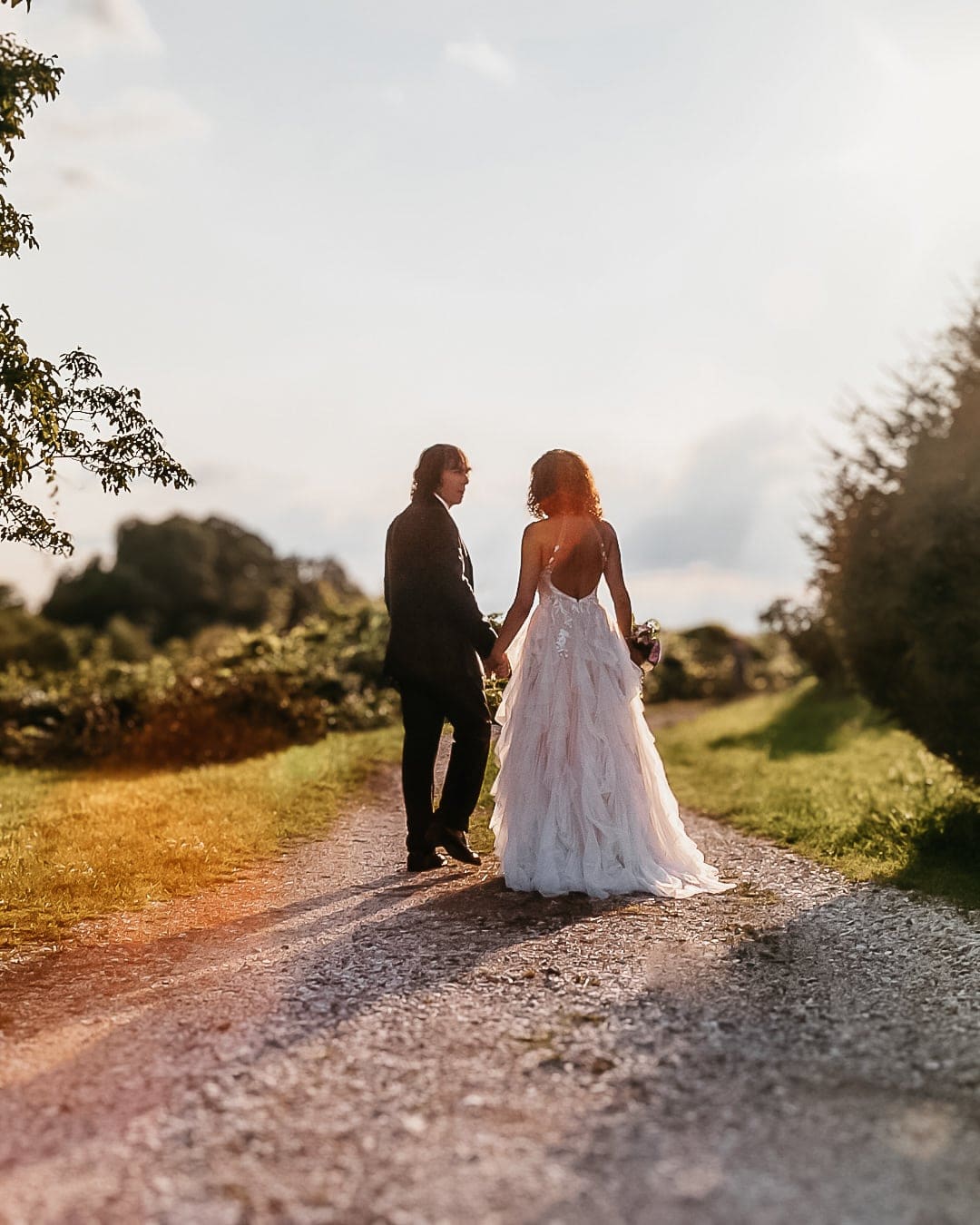 Sposi di spalle sul sentiero di campagna al tramonto, mano nella mano, con luce dorata e cielo chiaro, matrimonio a Pagnacco