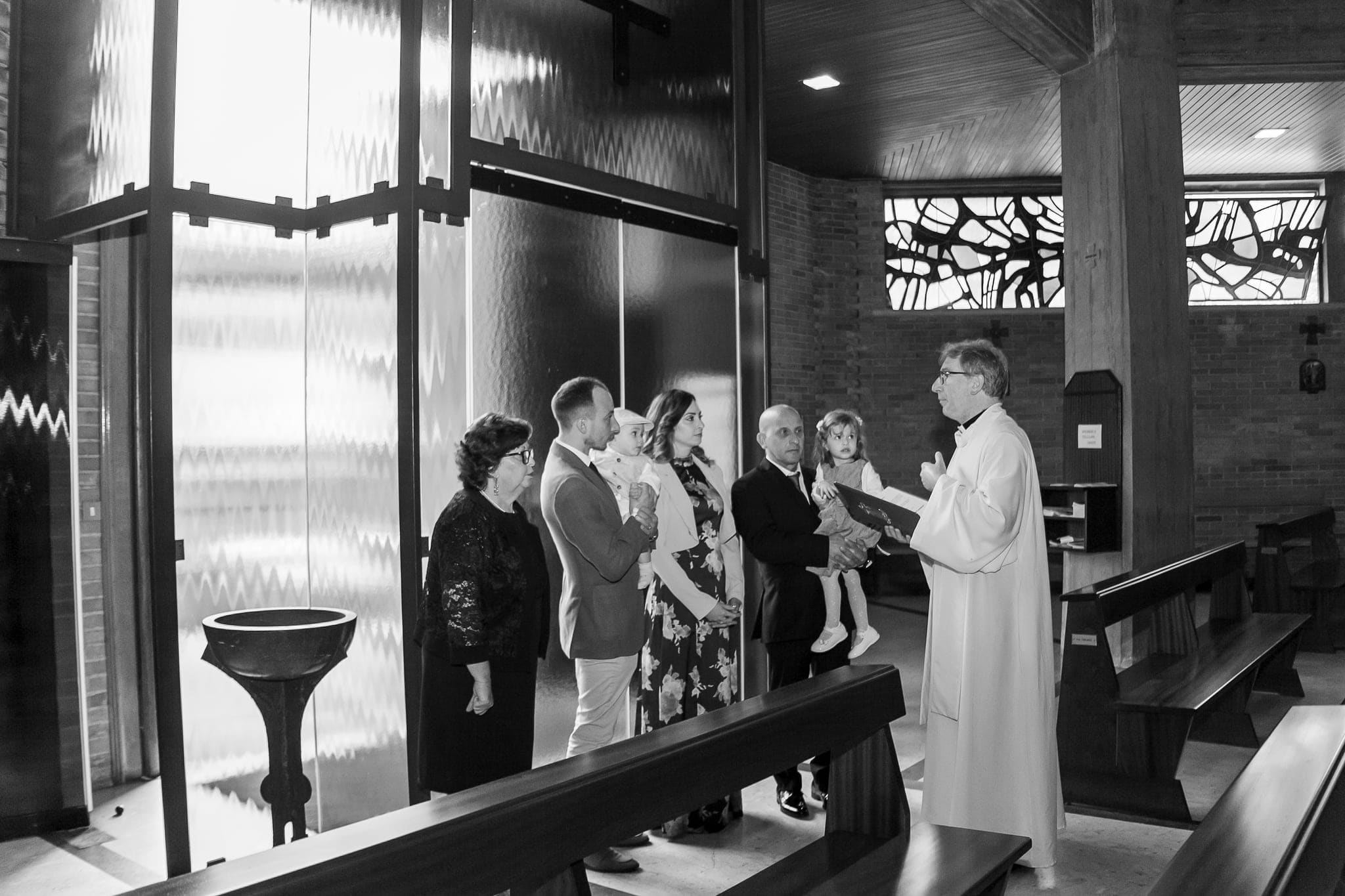 In bianco e nero, il sacerdote benedice la famiglia vicino al fonte, in una chiesa moderna con grandi vetrate