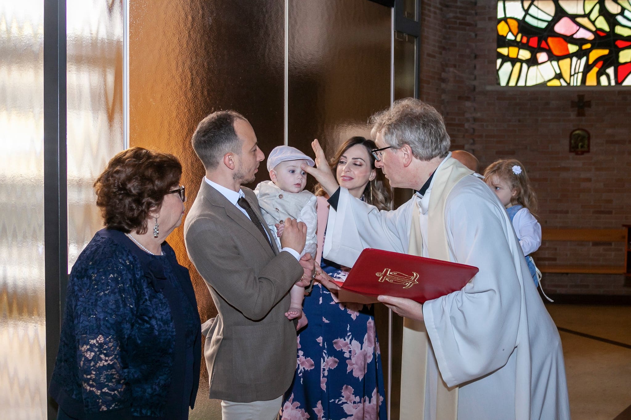 Il sacerdote con il libro rosso benedice il piccolo in braccio al papà, accanto alla mamma, a una nonna e alla sorellina