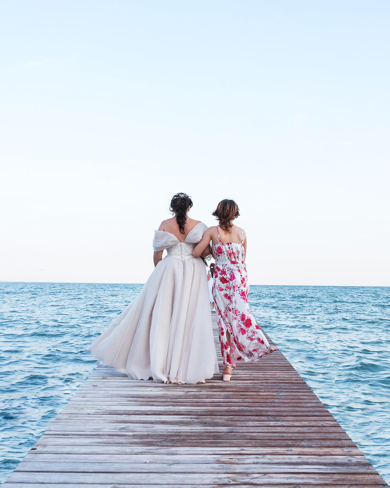 Sposa e un'amica con abito a fiori camminano di spalle sul molo in legno verso il mare aperto, matrimonio a Grado