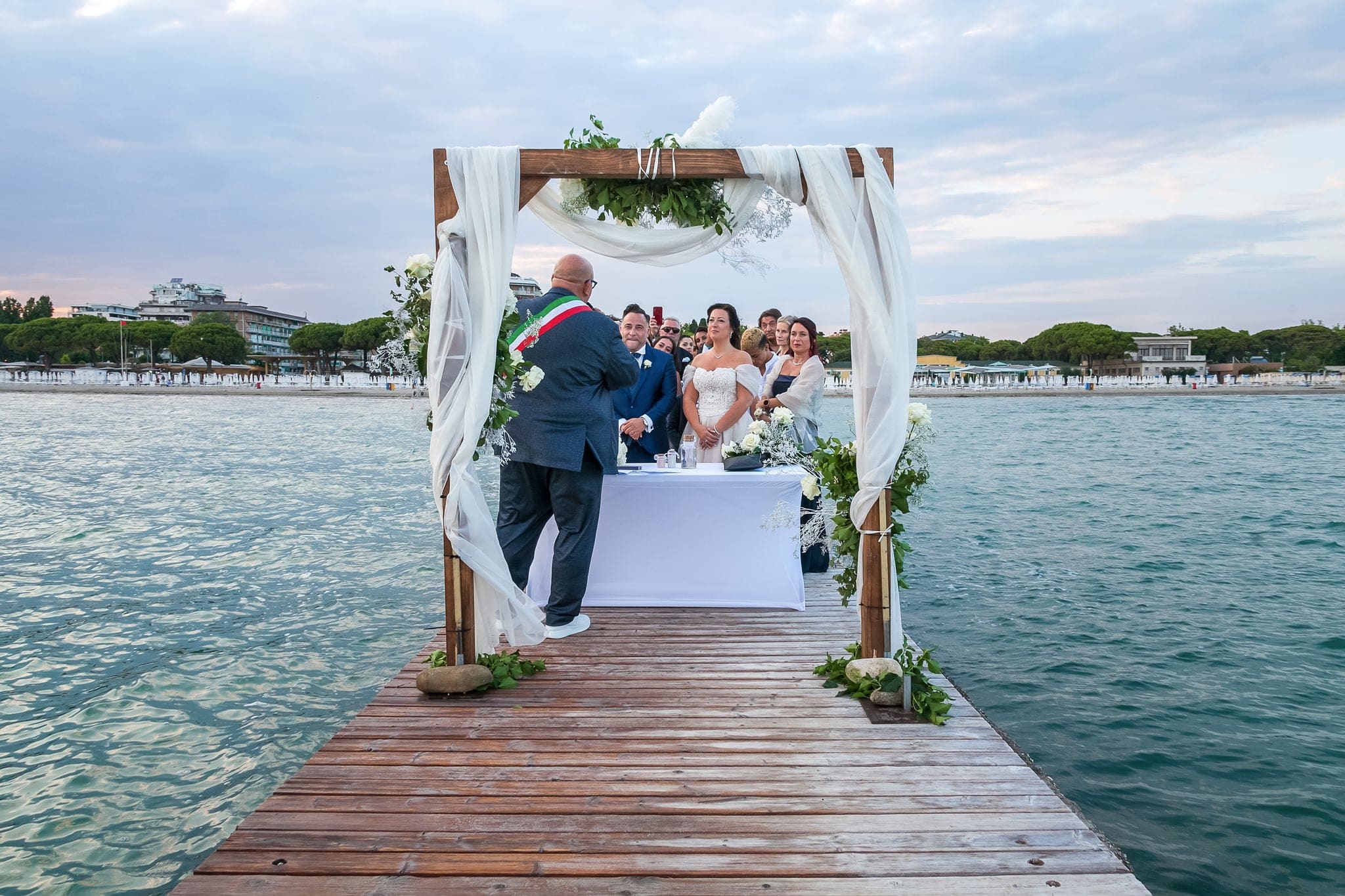 Cerimonia civile su un molo in legno con arco decorato di tessuto e foglie, celebrante e sposi davanti al mare di Grado